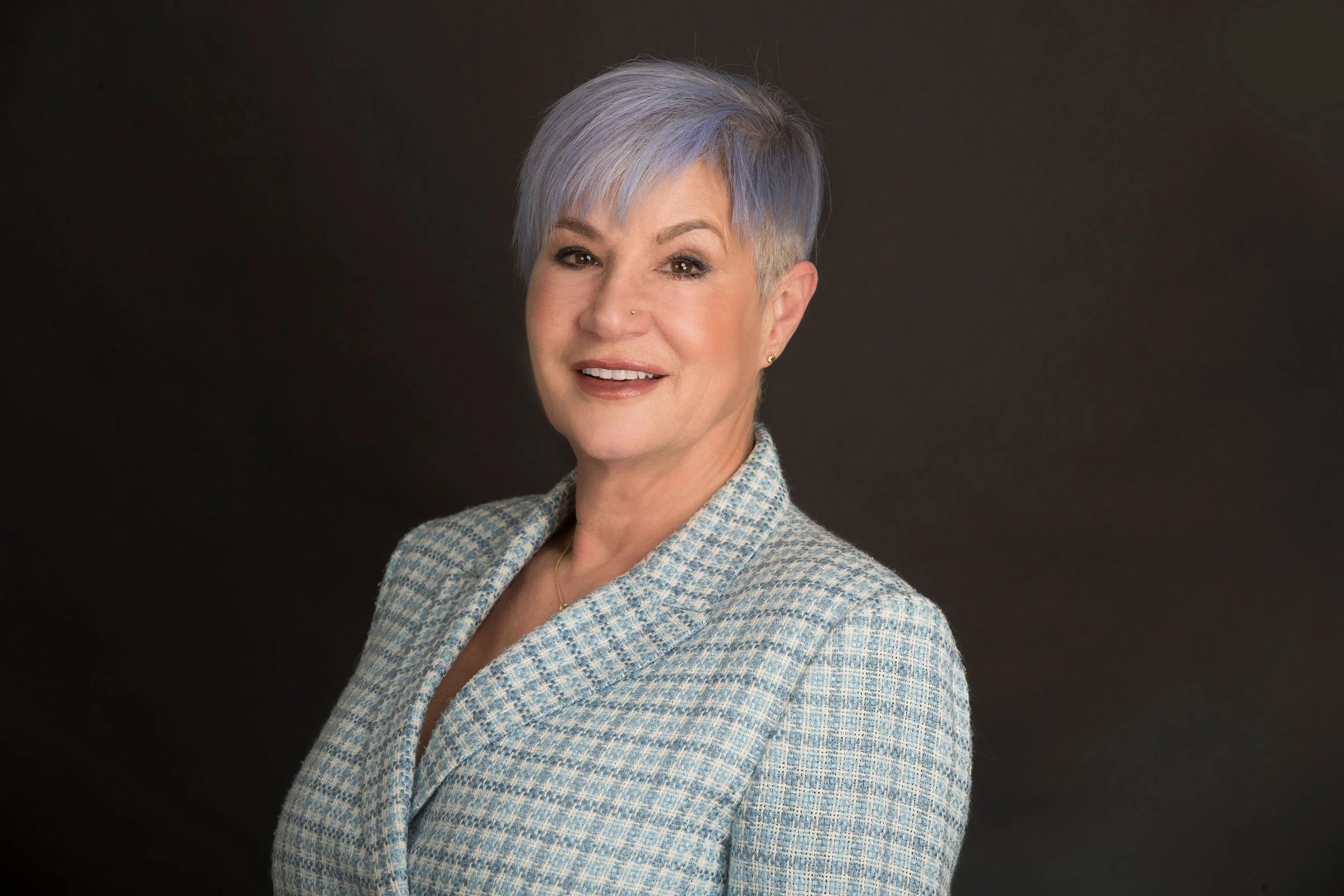 A portrait of a woman with short, styled purple hair, smiling, wearing a patterned blazer, against a dark background.