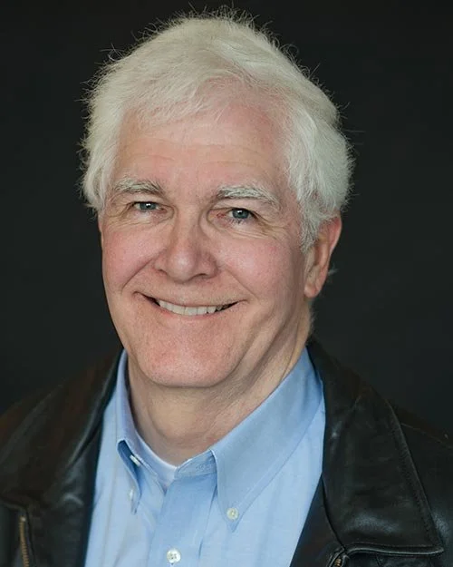Portrait of an older man with white hair, smiling, wearing a light blue shirt and a black leather jacket, against a dark background.