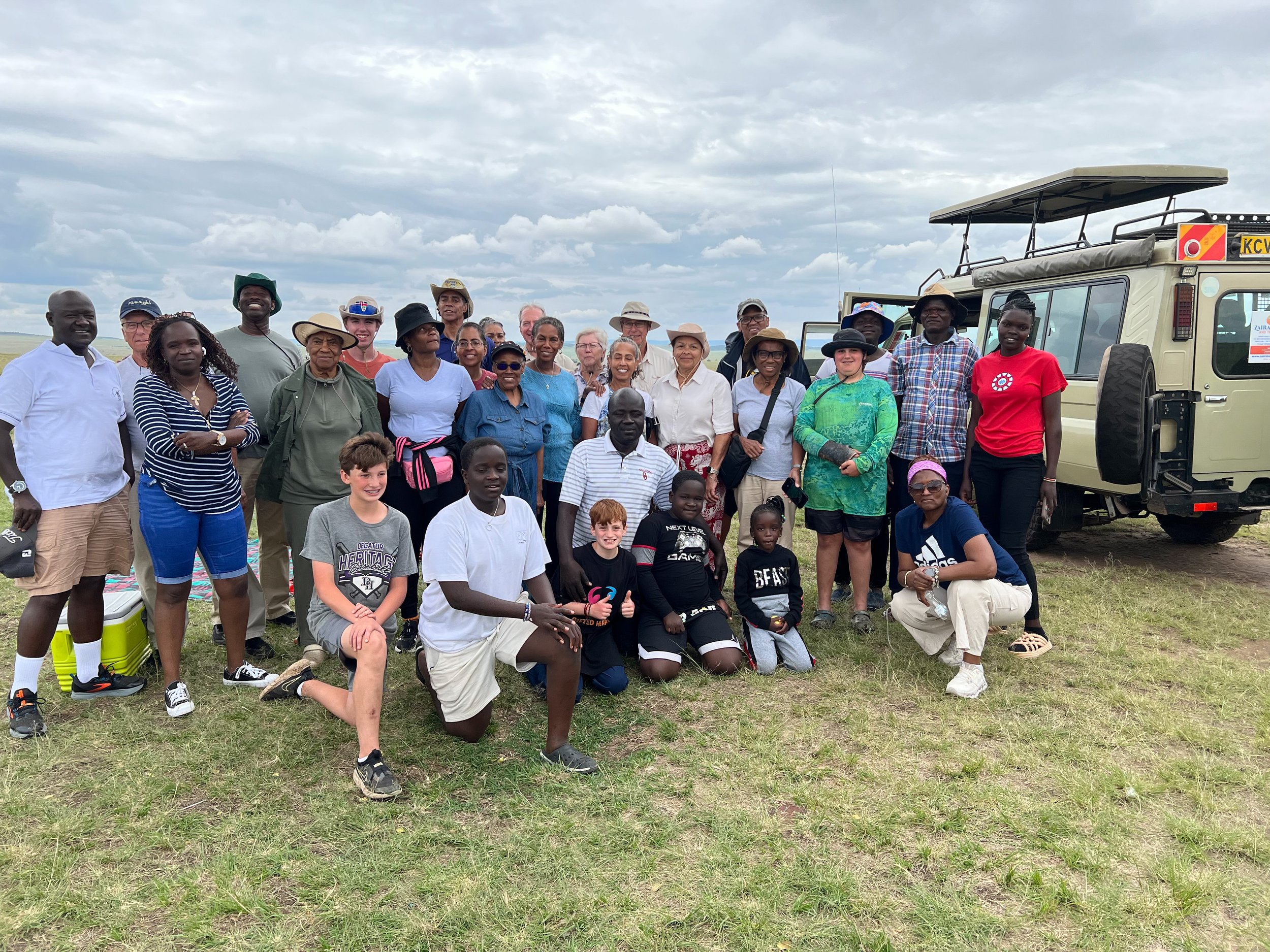 A group of diverse people posing together outdoors near a safari vehicle. The group consists of adults and children, some kneeling and some standing, dressed in casual outdoor attire. In the background is a cloudy sky and a grassy area typical of a s