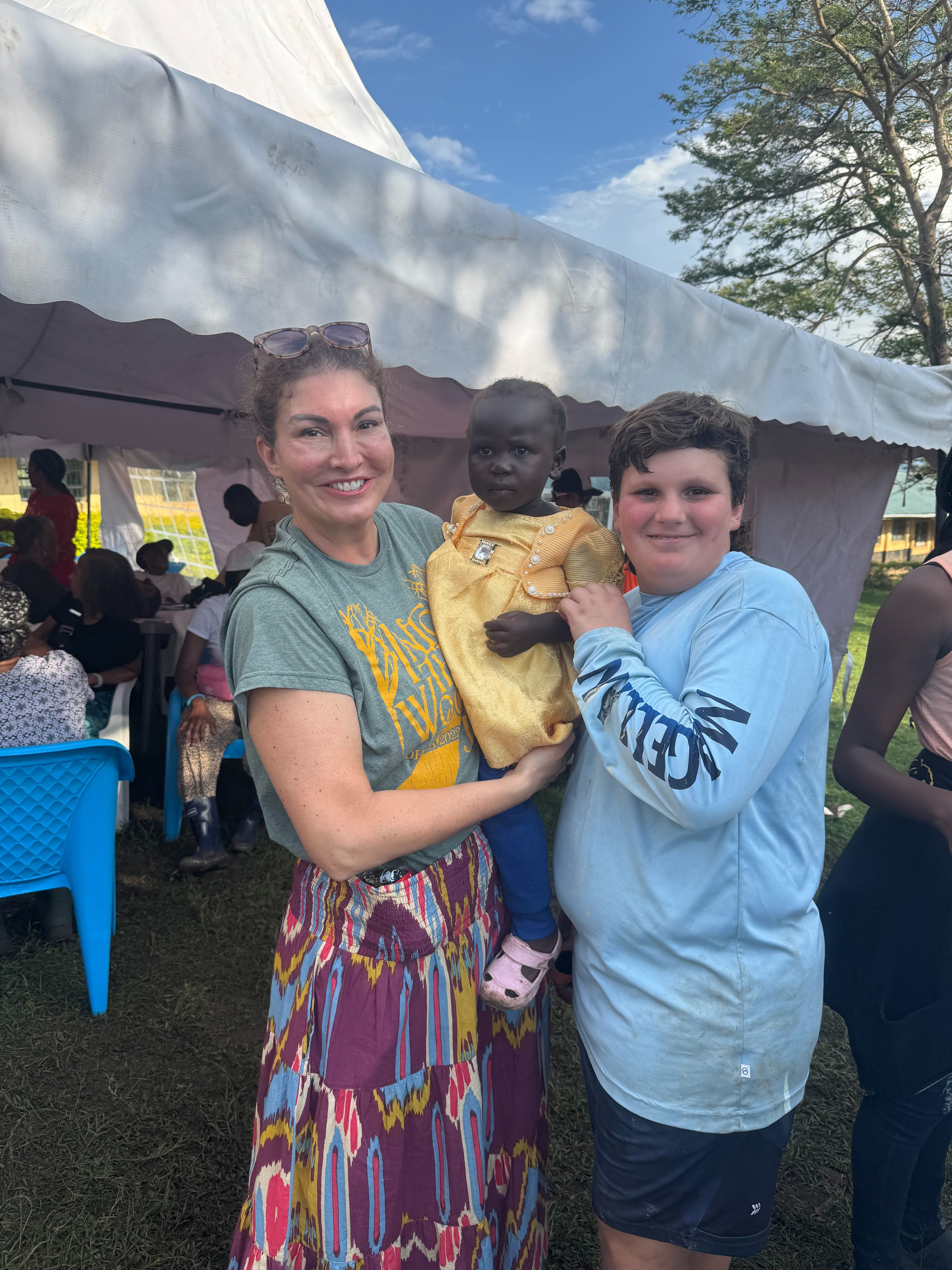 A woman and a boy hold a young child in their arms, smiling at the camera. They are standing outdoors near a white tent with people seated under it. The woman is wearing a green shirt and colorful skirt, while the boy is wearing a light blue long sle