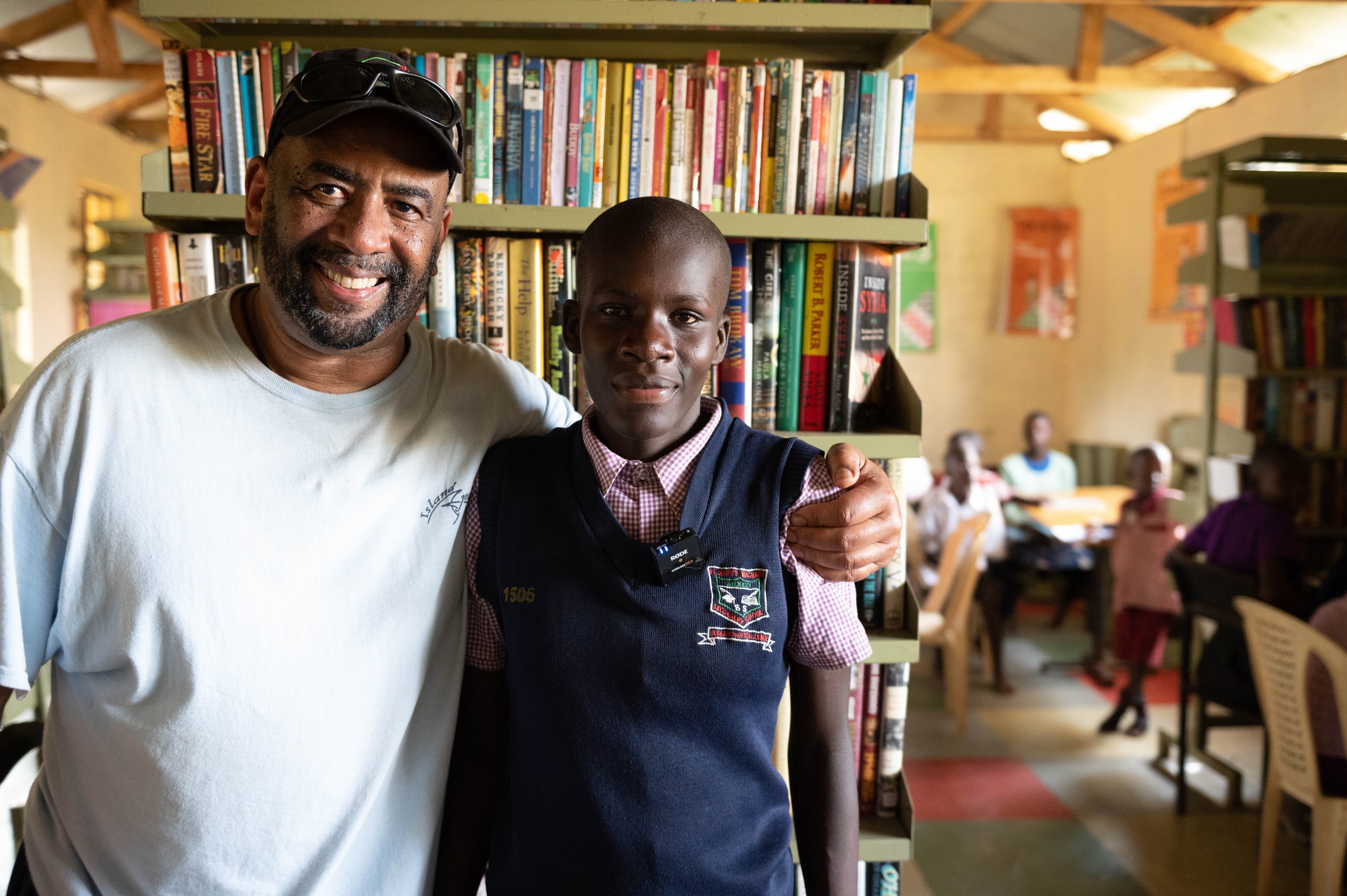 Man and teenager smiling in a library with bookshelves in the background.