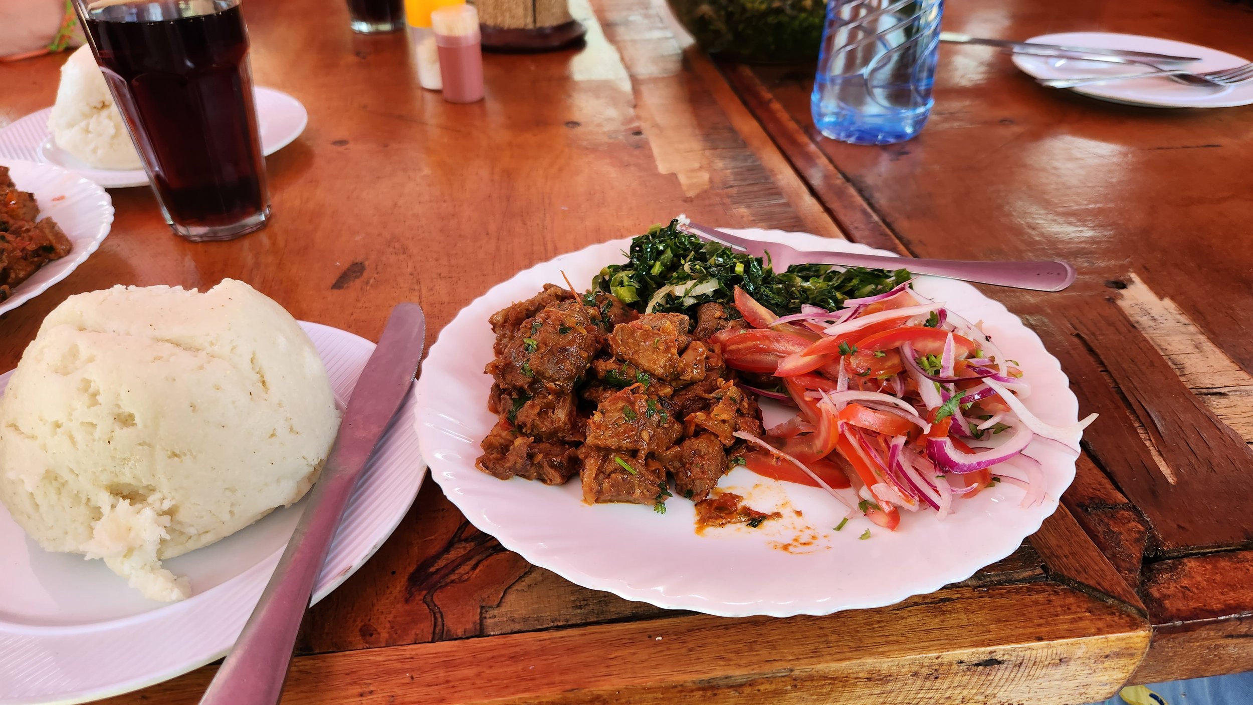 Plate of food with cooked meat, greens, sliced tomatoes and onions, and a separate plate with a serving of ugali on a wooden table.