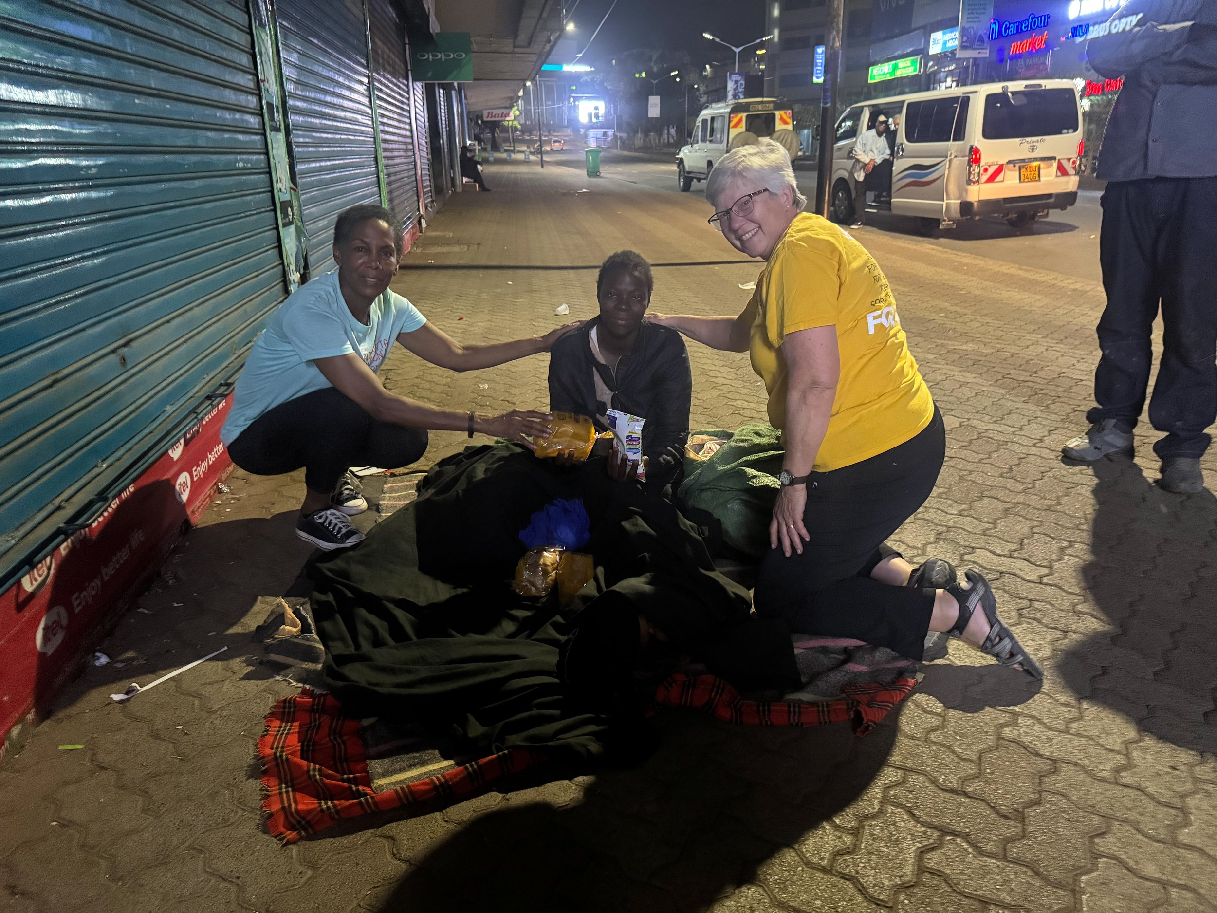 Two women kneel beside a seated person on a sidewalk, offering food items. The scene takes place at night in an urban area with view of closed stores and parked vehicles.