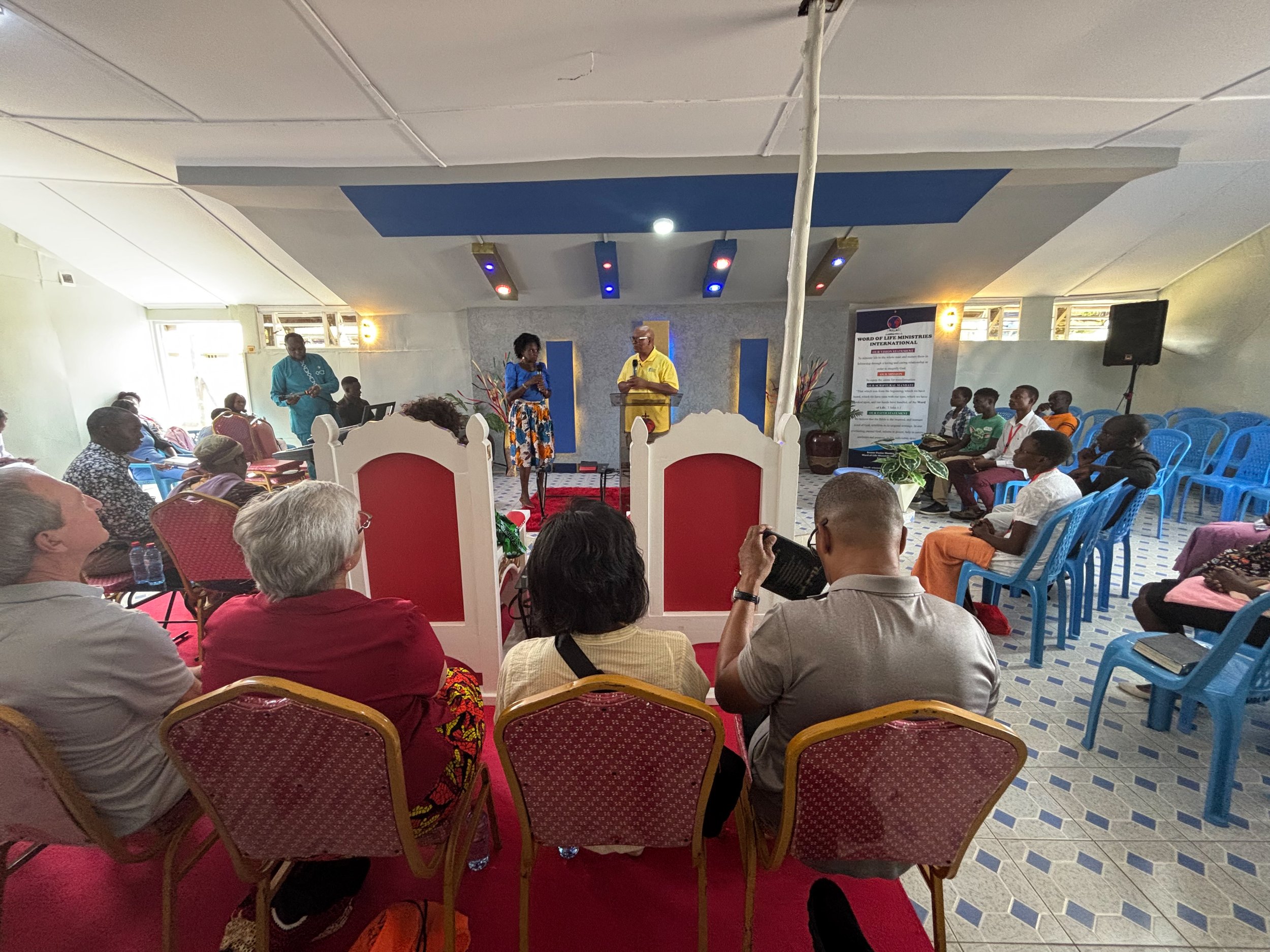 Indoor gathering with people seated on chairs, a speaker on stage, and a banner in the background.