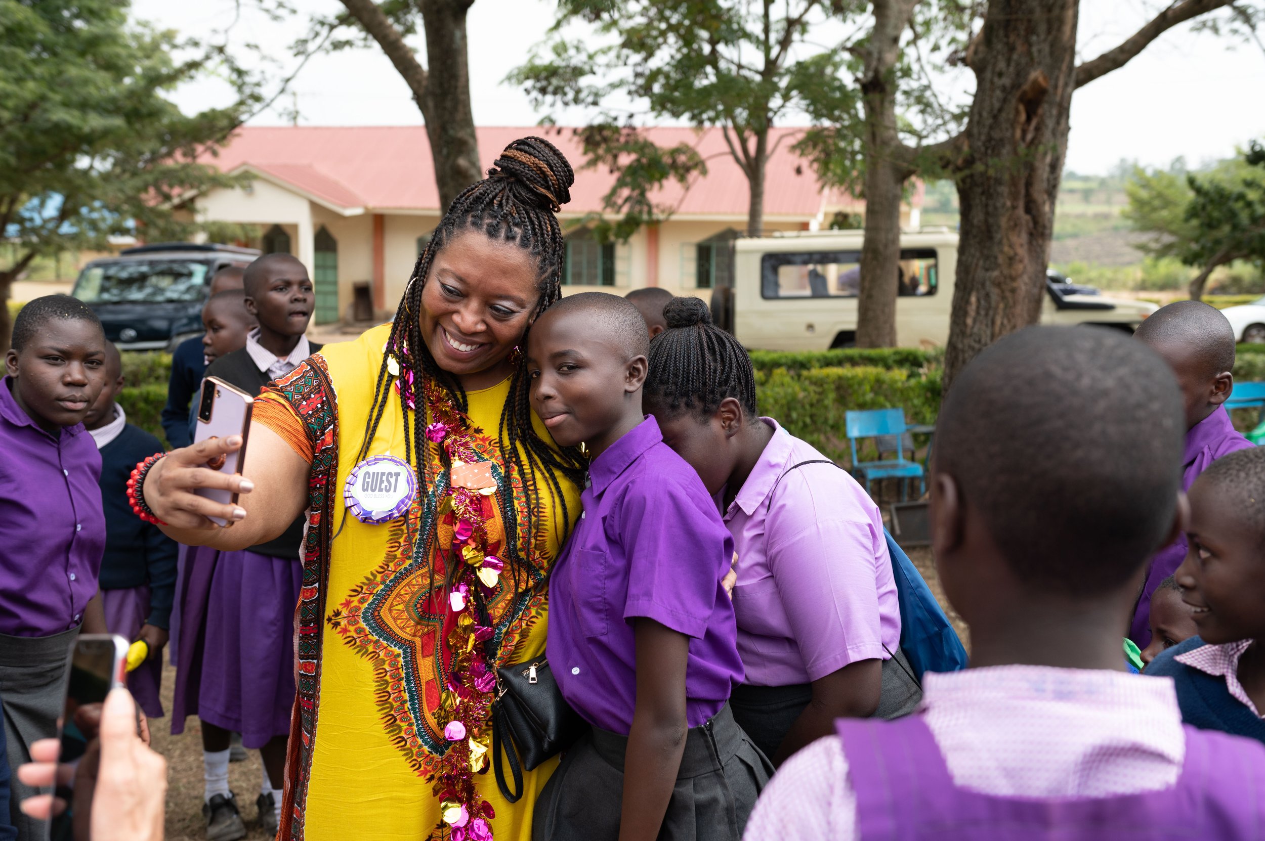 A woman in a yellow dress with a 'guest' badge takes a selfie with a group of students in purple uniforms outdoors. Trees, a vehicle, and a building are visible in the background.