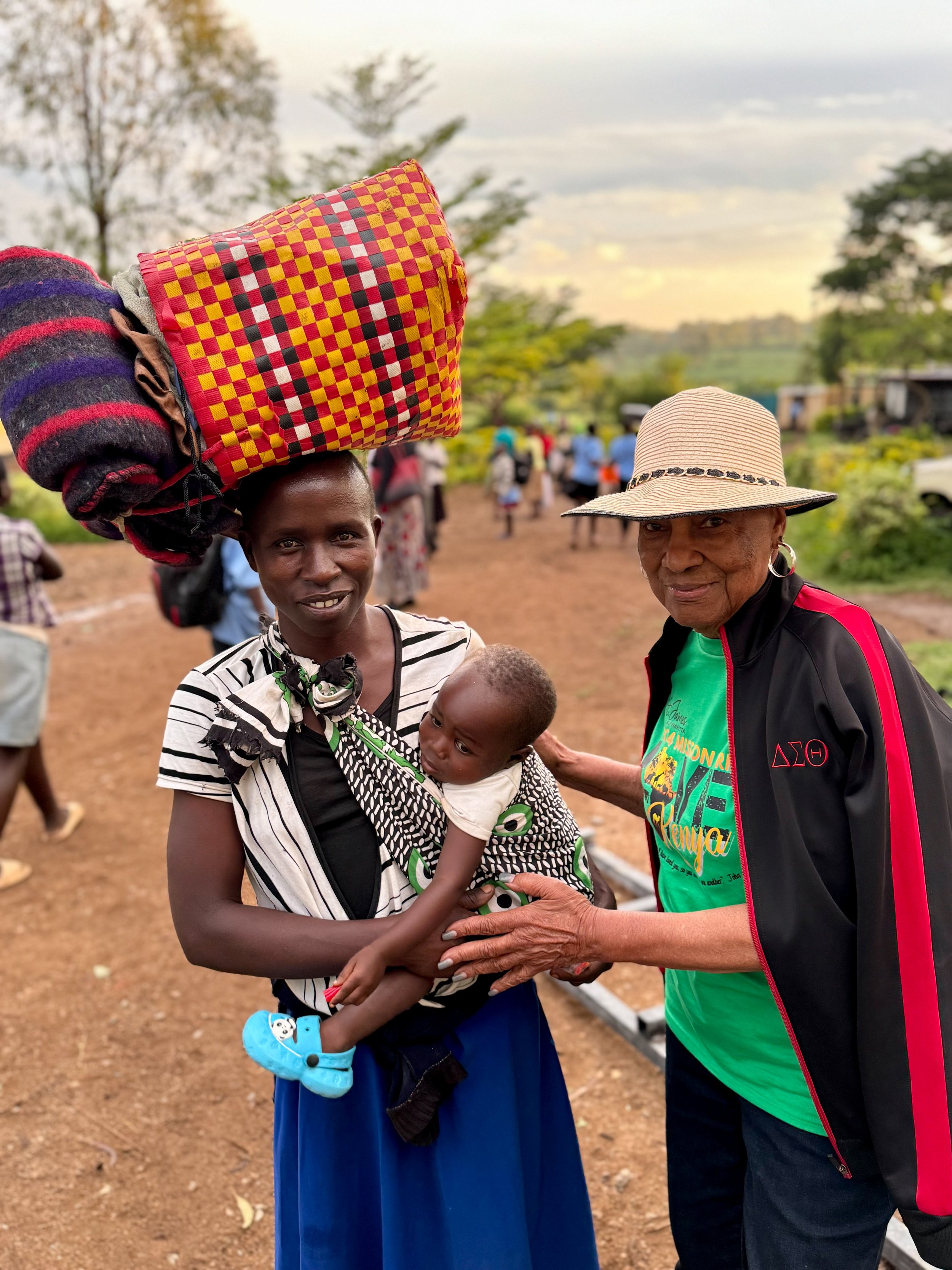 Two people, one holding a baby, standing outdoors. The person on the left balances a bundle on their head and carries a baby in a patterned wrap. The other person wears a hat and jacket. A rural setting with trees and people in the background.