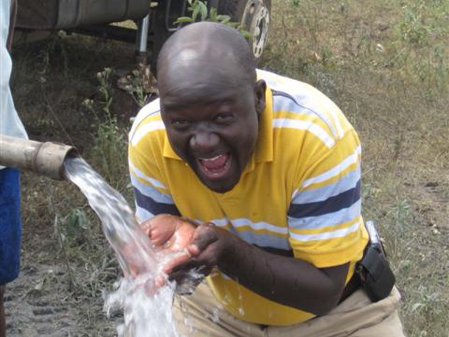 A man in a yellow striped shirt is holding water in his hands, with water pouring from a pipe onto his hands. He appears to be reacting with surprise and excitement, outdoors on a grassy area.