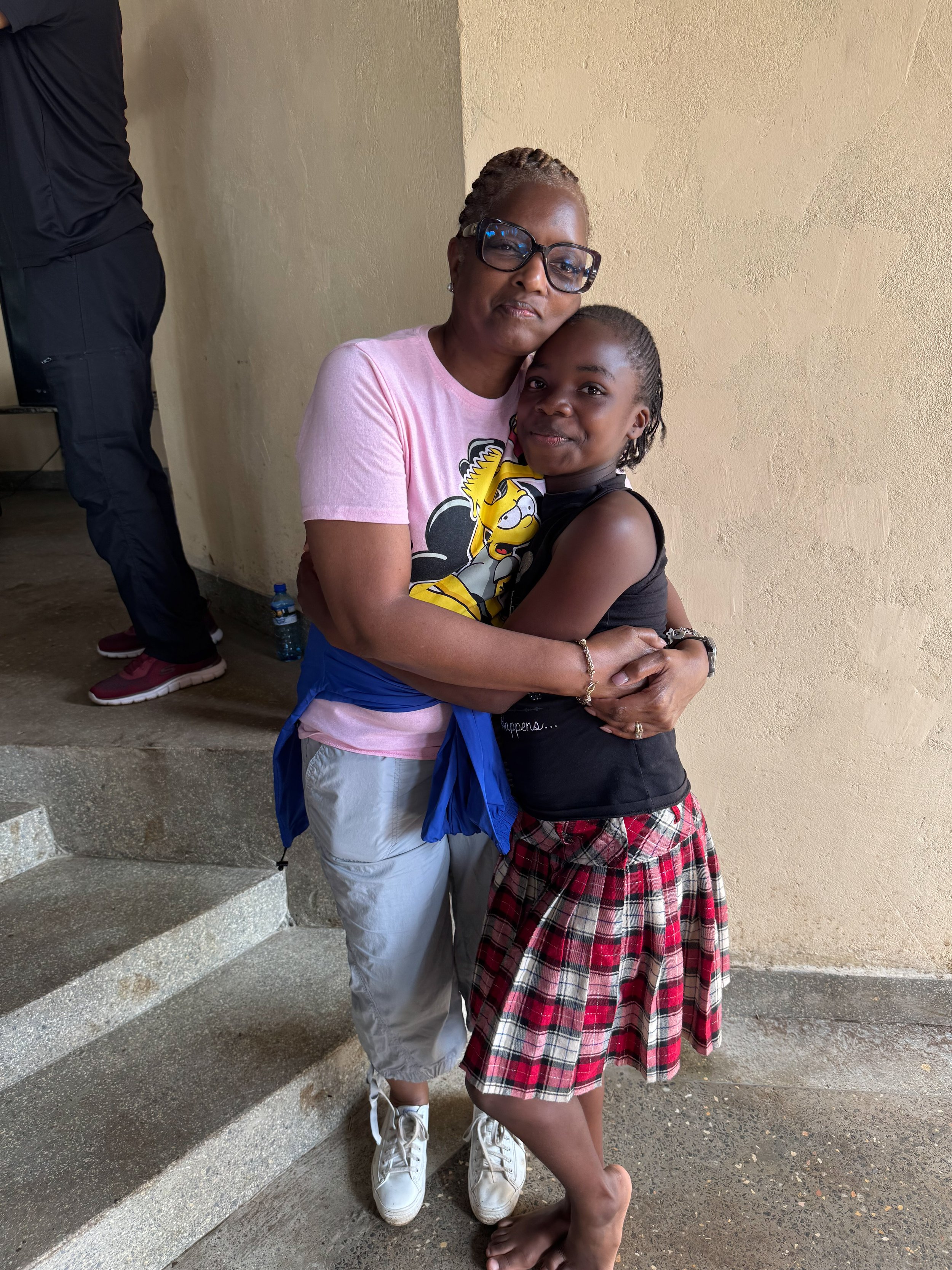 A woman in a pink T-shirt is hugging a young girl in a black top and red plaid skirt. They are standing on a staircase indoors, with the woman wearing white sneakers and glasses.