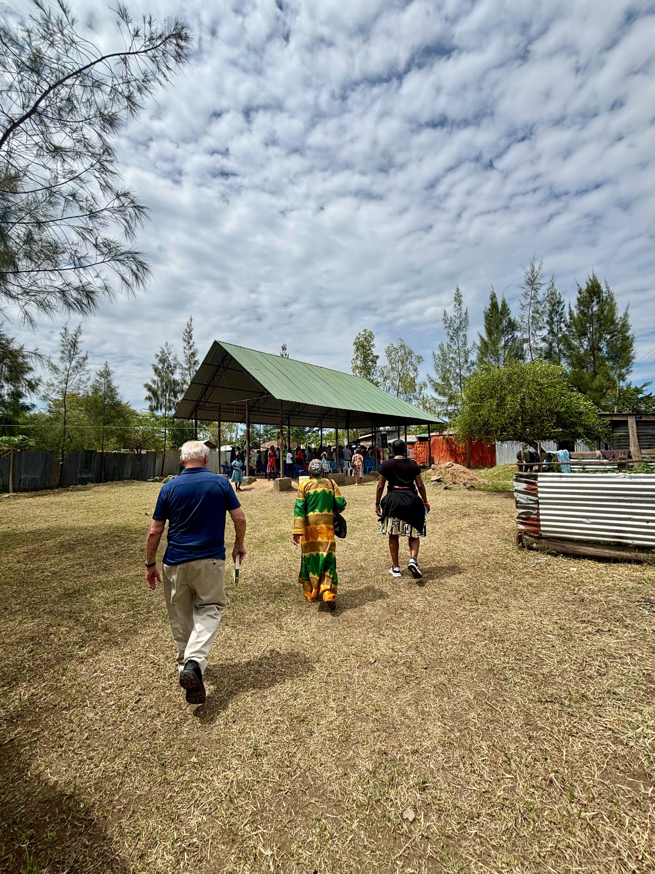 People walking towards a large tent in an outdoor setting with trees and a cloudy sky.