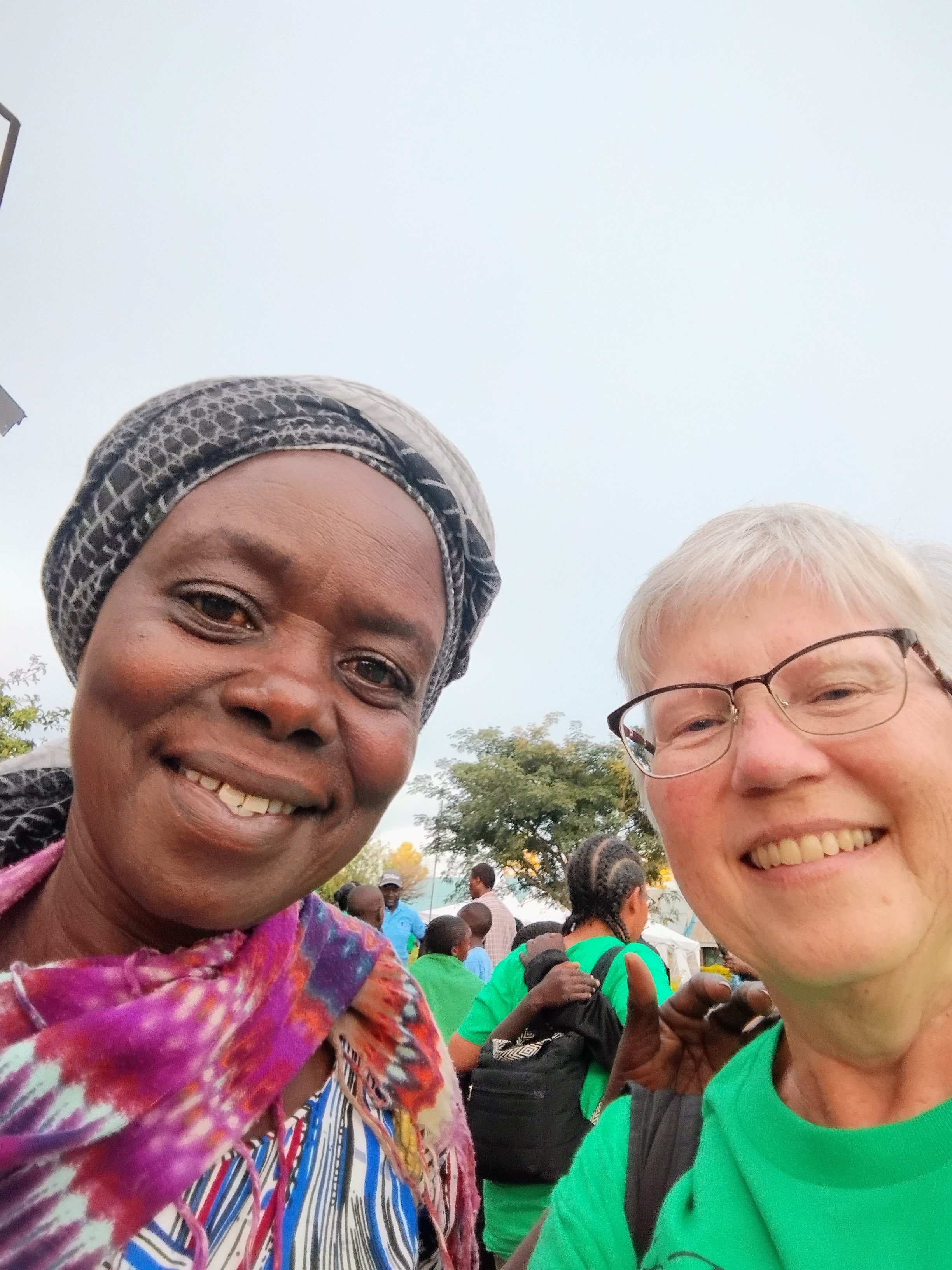 Two smiling women taking a selfie outdoors, surrounded by people wearing green shirts.
