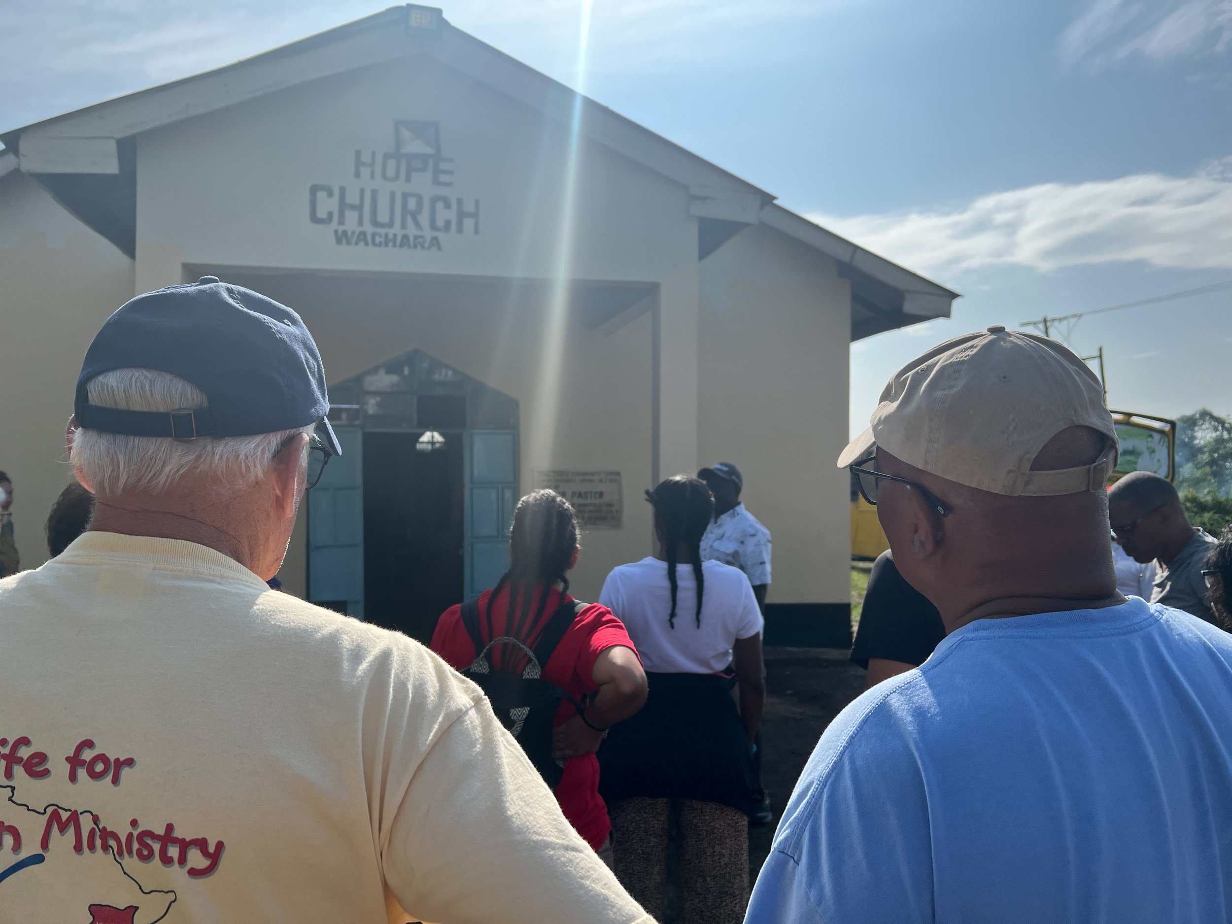 Group of people standing outside Hope Church Wachara, with two men in the foreground wearing hats and casual clothing, looking towards the church entrance.