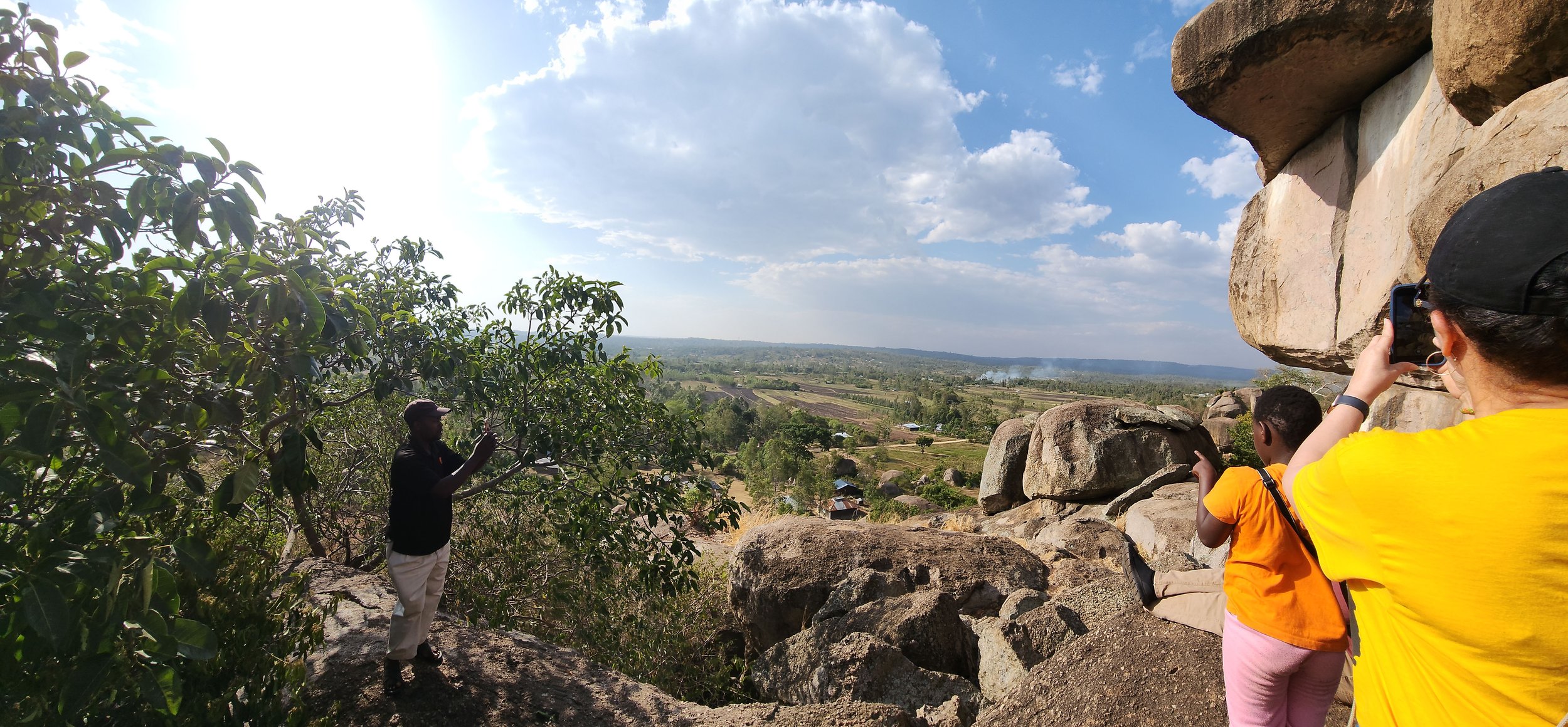 People taking photos of a scenic landscape view with rocks, trees, and wide sky, overlooking a rural area with distant fields and smoke rising.