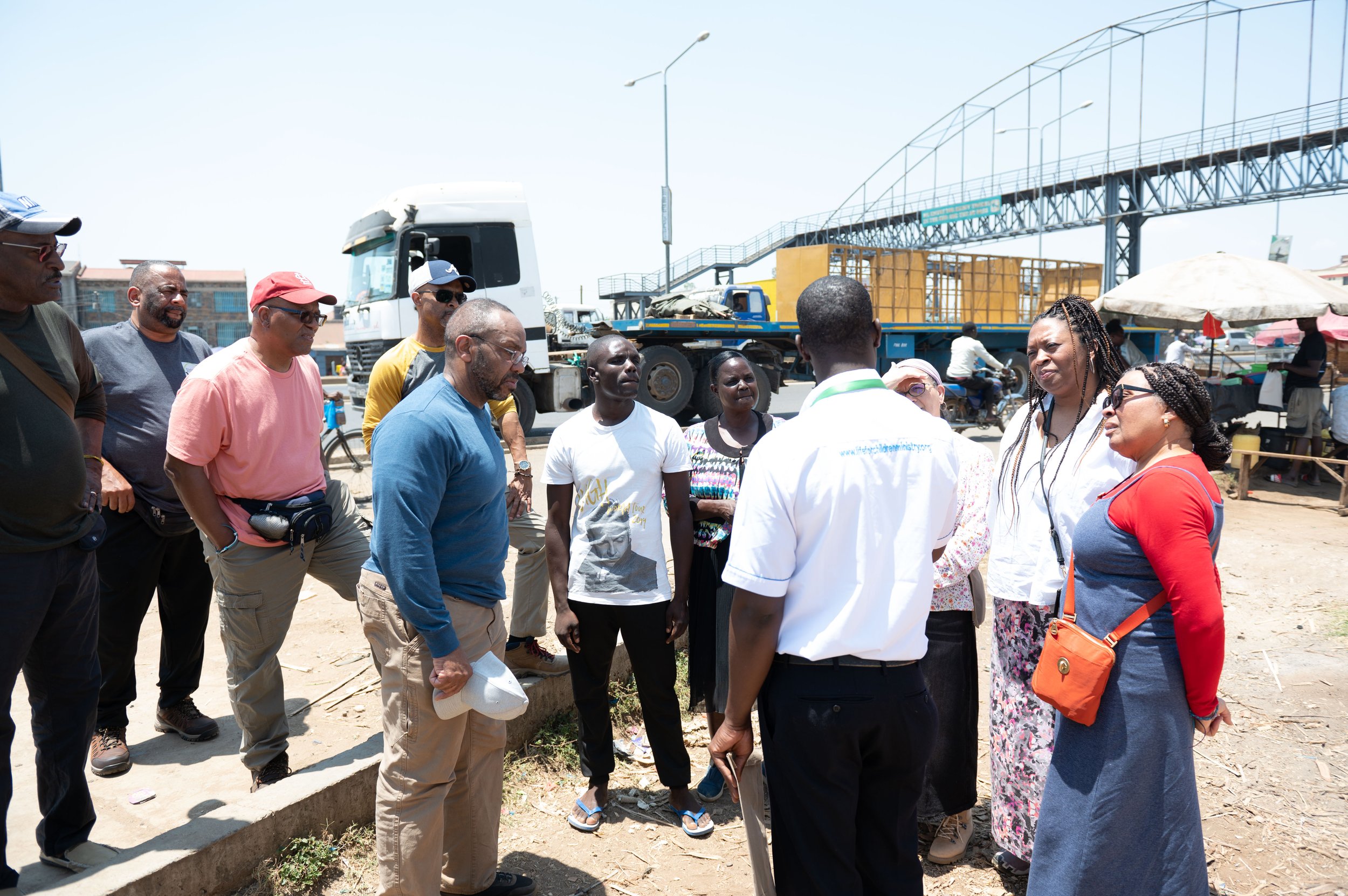 A group of people gathered outdoors near a busy street with trucks parked in the background, under a clear sky.