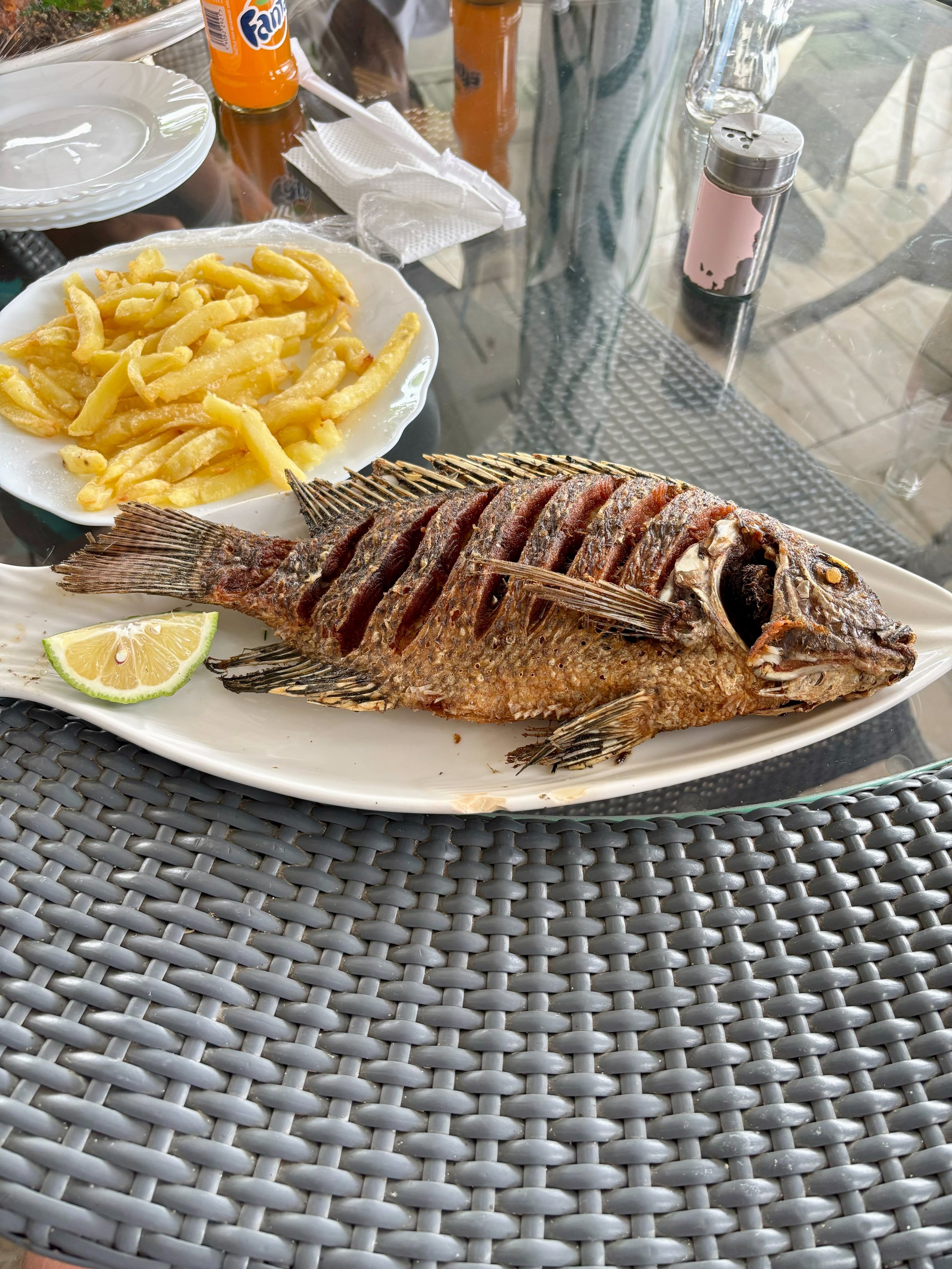 Fried whole fish with lime slice on a white plate alongside a plate of fries on a glass table.