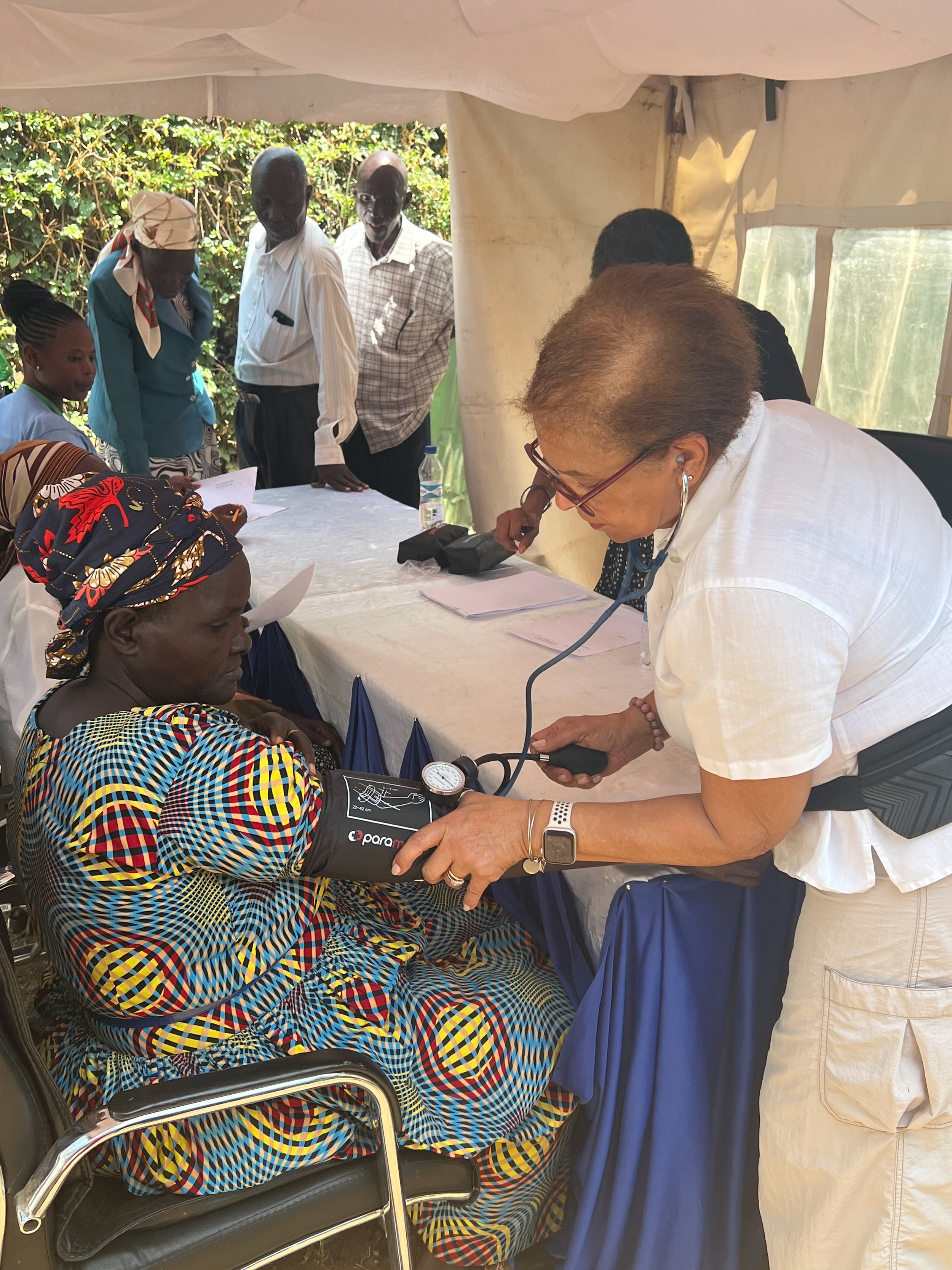A healthcare worker is taking a woman's blood pressure in a makeshift clinic setting. The woman is seated and wearing a colorful patterned dress. Several other people are in the background, some standing and others sitting.