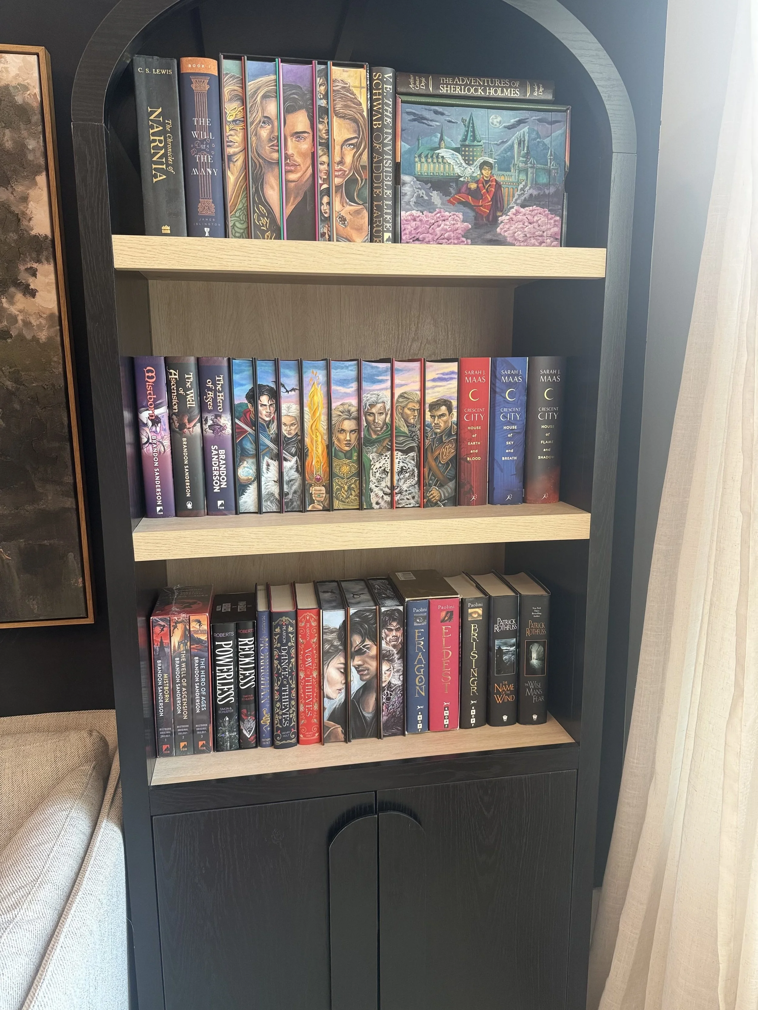 A bookshelf with three shelves filled with books. The top shelf has a set of hardcover books, a colorful painting, and a book titled 'The Adventures of Sherlock Holmes.' The middle shelf features a series of fantasy novels by Brandon Sanderson, including 'Mistborn,' 'The Well of Ascension,' and 'The Hero of Ages.' The bottom shelf contains more books, including some about power and dragons, with titles such as 'Power and Dragons' and 'The Wise Man's Fear.' The bookshelf is black with a natural wood-colored interior.
