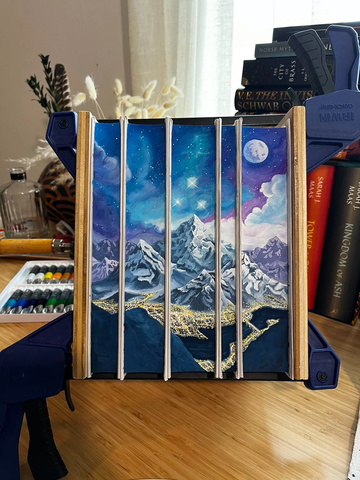 A metal art piece depicting a mountain landscape under a starry night sky with the moon, set inside a wooden frame and placed on a blue easel, surrounded by books and art supplies.