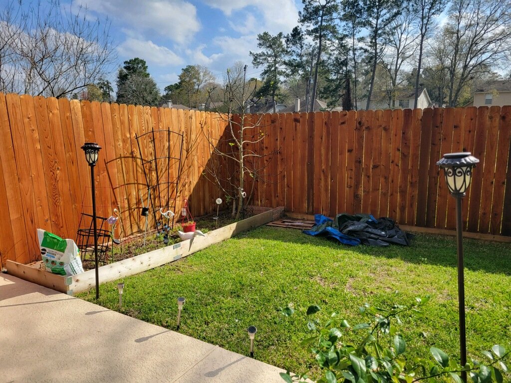 Backyard with a wooden fence, patio area, garden bed with potted plants, outdoor lights, and a tarp on the grass.