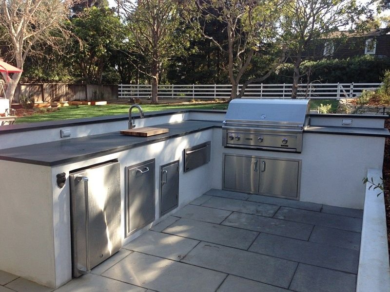 Outdoor kitchen with stainless steel appliances, grill, and countertop, set in a backyard with trees and fencing.