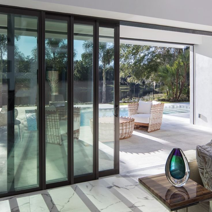 Sliding glass doors separating a modern living room with a marble floor from an outdoor patio area featuring wicker furniture, a pool, and surrounding trees.