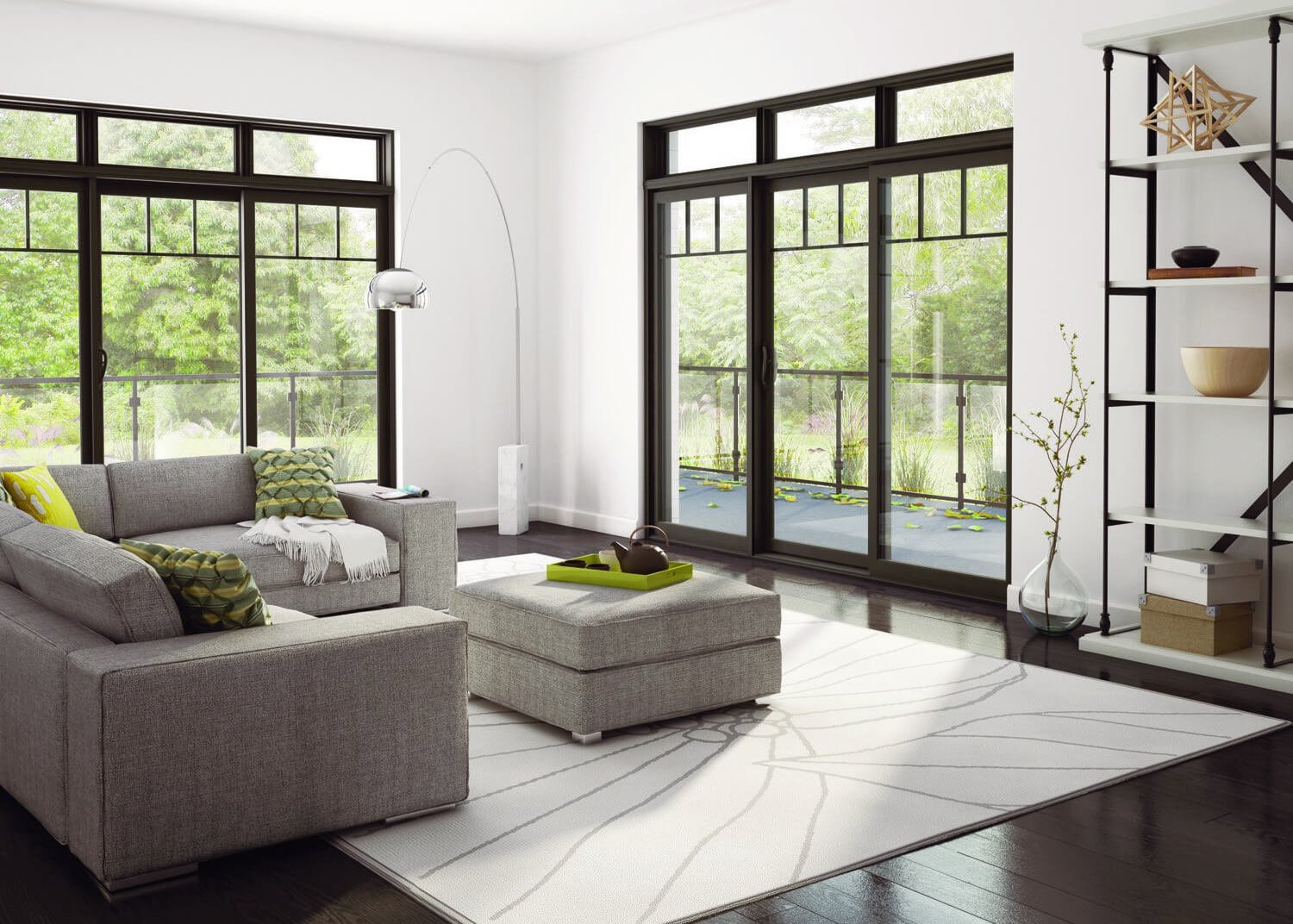 Modern living room with large windows, gray sectional sofa, ottoman, and black metal bookshelf. Green throw pillows and a white rug are present. Outdoor greenery visible through glass doors.