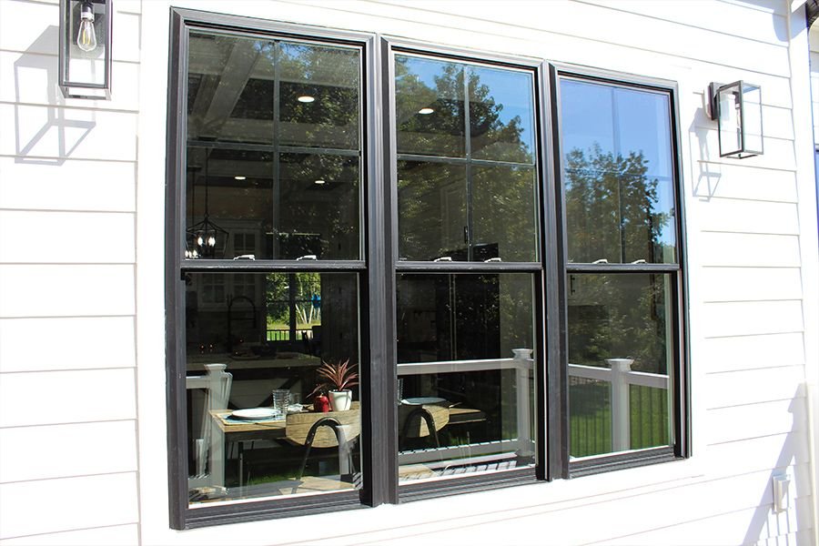 Three large black-framed windows on a white house exterior with reflections of trees and an outdoor seating area.