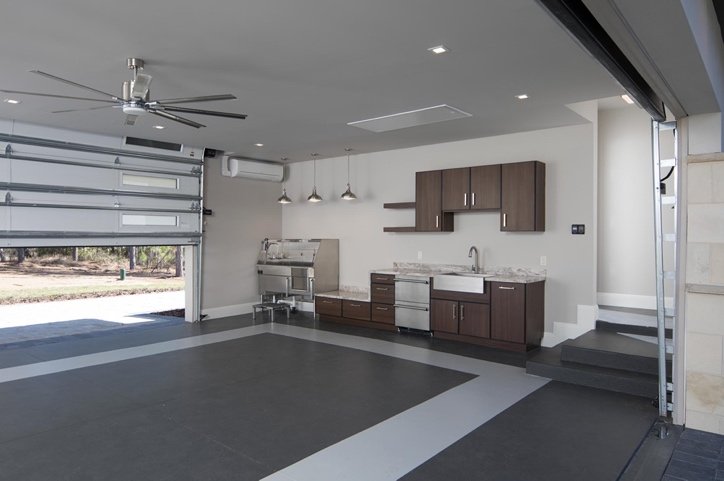 Modern garage interior with a ceiling fan, open garage door, and a small kitchenette area with dark wood cabinets, stainless steel appliances, and marble countertops.