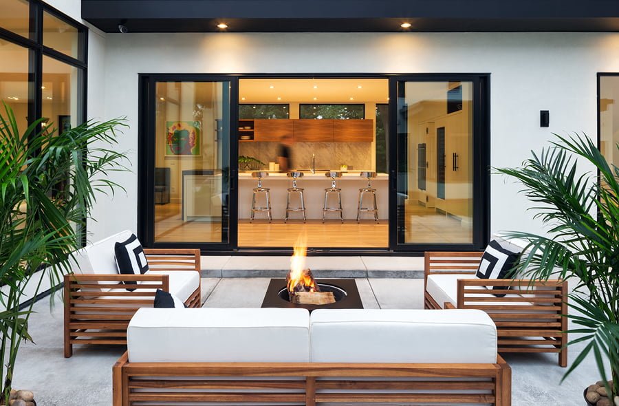 Modern outdoor patio with a fire pit, white cushioned wooden seating, and tropical plants. Sliding glass doors reveal a kitchen with bar stools inside.
