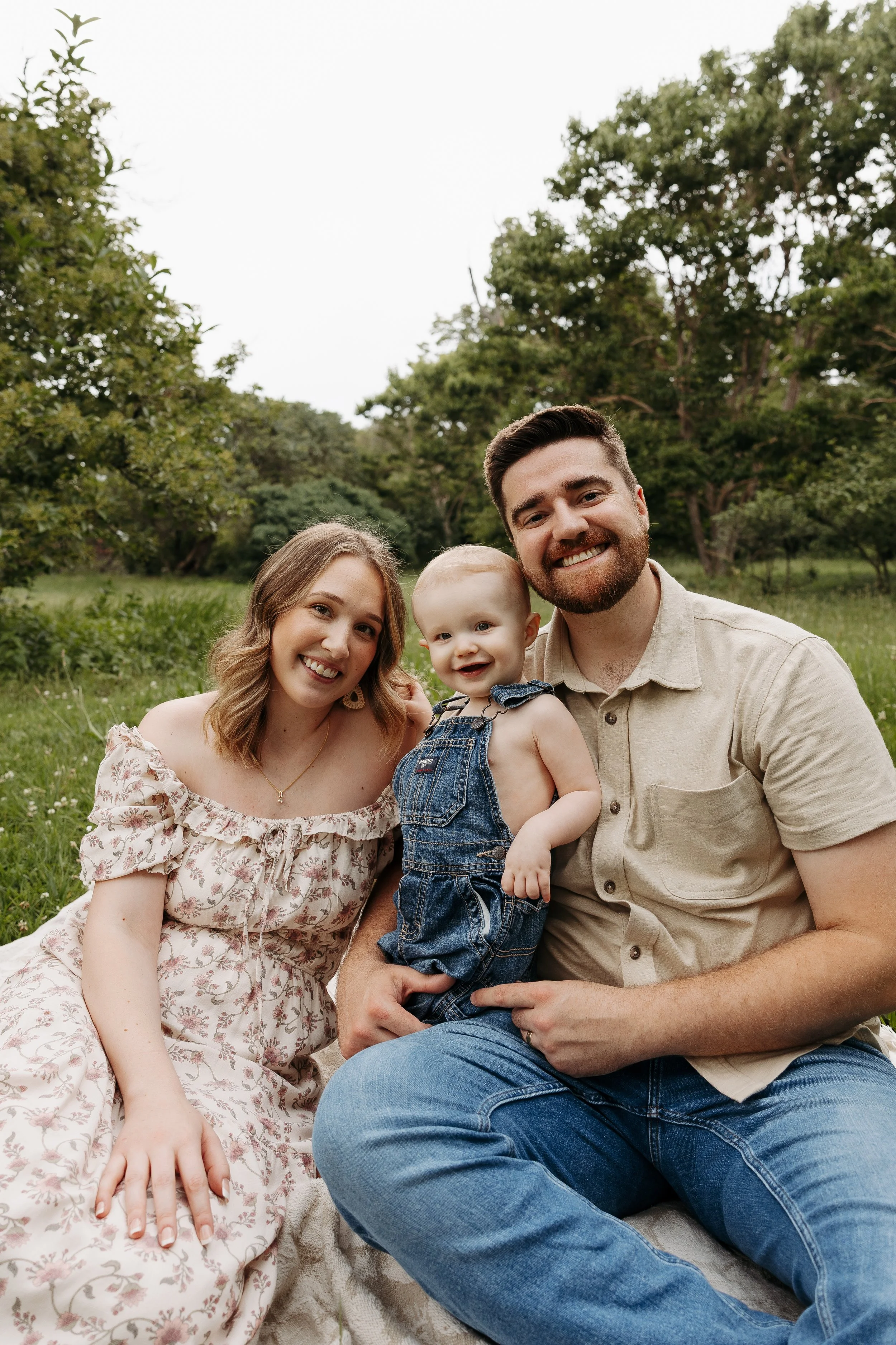 A happy family of three sitting outdoors in a grassy area with trees in the background. The family consists of a woman with long, wavy blonde hair, a man with short brown hair and a beard, and a young child with short blonde hair. The woman is wearing a light-colored floral off-shoulder dress, the man is wearing a beige shirt, and the child is dressed in denim overalls.