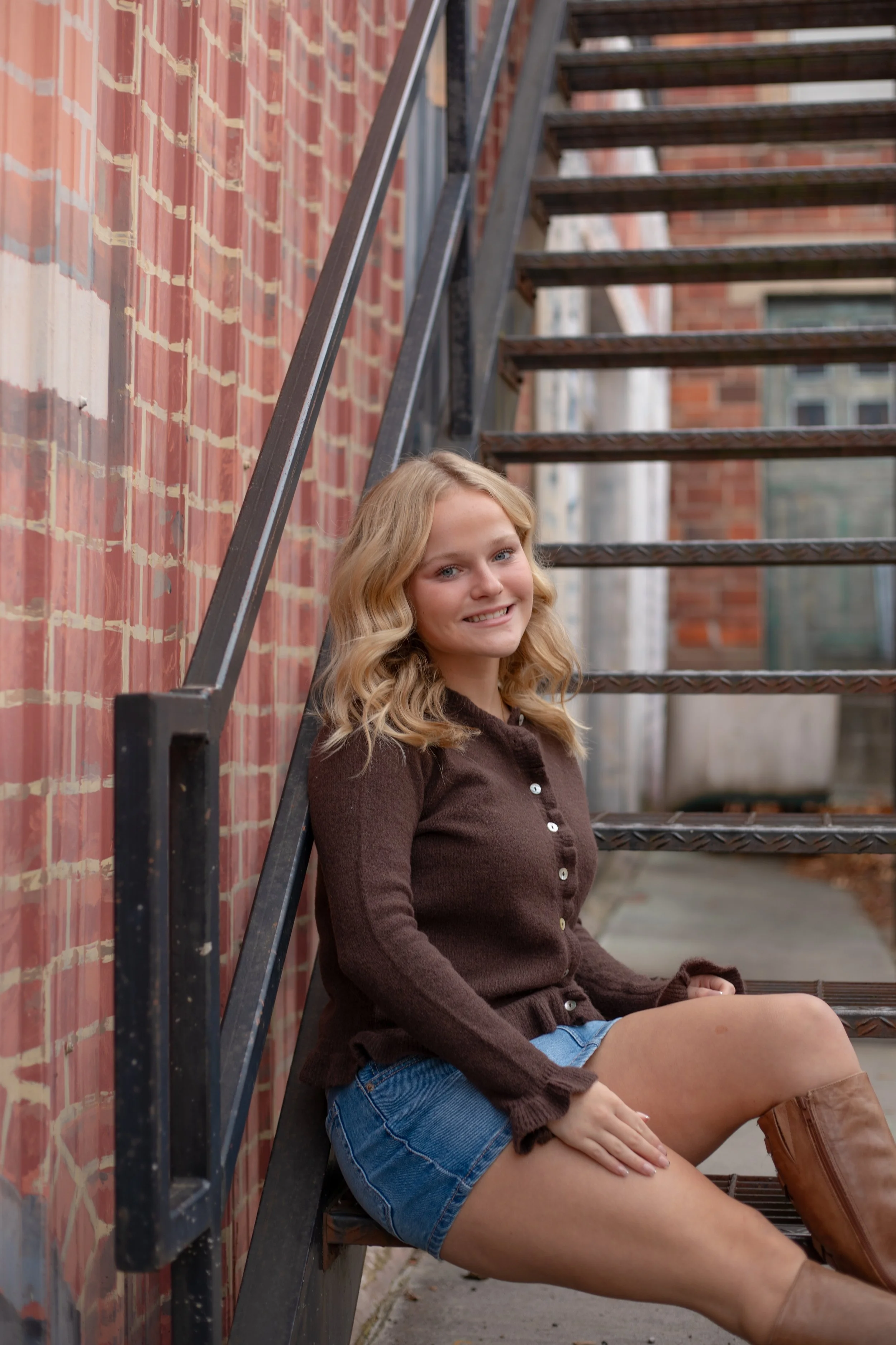A young woman with blonde wavy hair, wearing a brown sweater, denim shorts, and brown boots, sitting on a fire escape ladder outside a brick building, smiling at the camera.