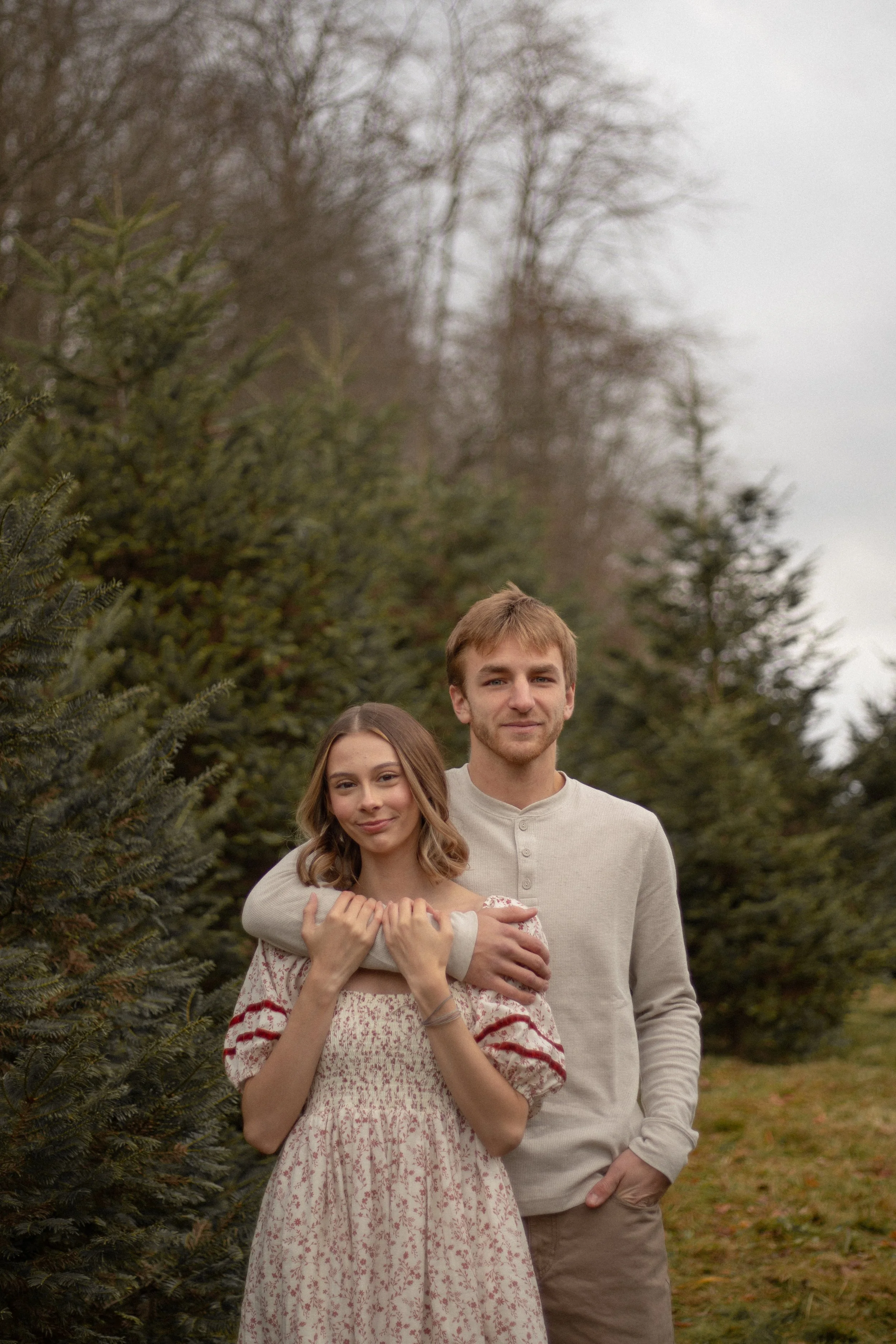 A young couple standing outdoors in front of evergreen trees during daytime, the woman is wearing a floral dress and has light brown hair, and the man is wearing a beige shirt and has short blonde hair.
