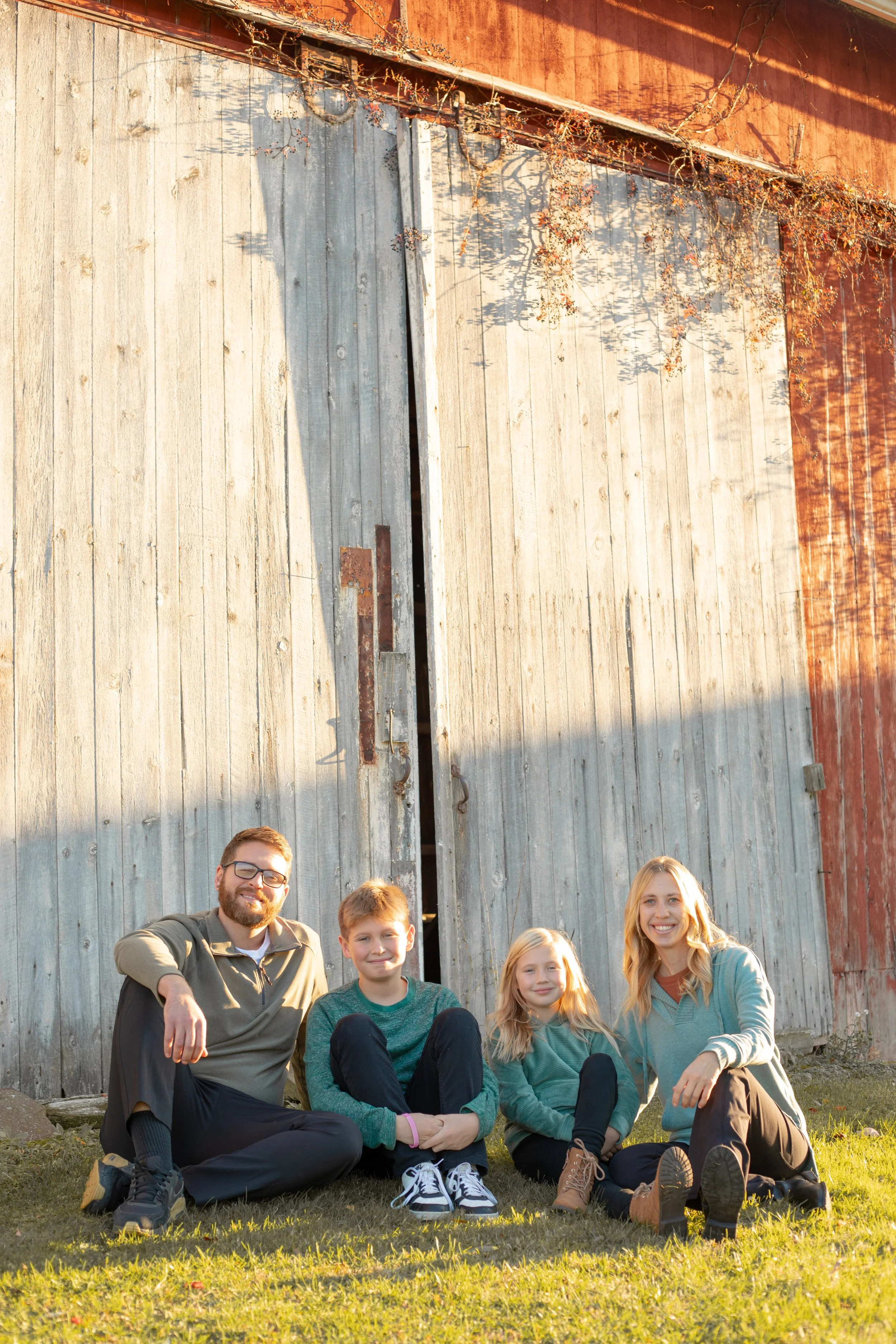 A family of four sitting on the grass in front of a rustic, weathered wooden barn with vines hanging on its upper part, during late afternoon sunlight.