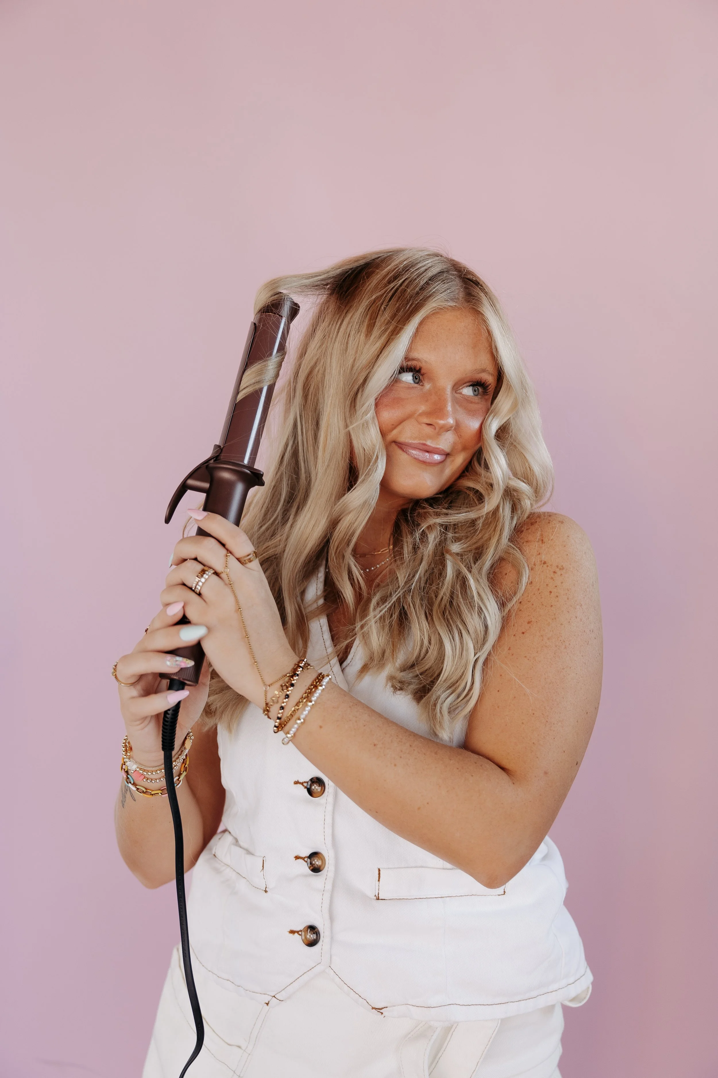 A woman with long blonde hair using a curling iron, standing against a pink background.