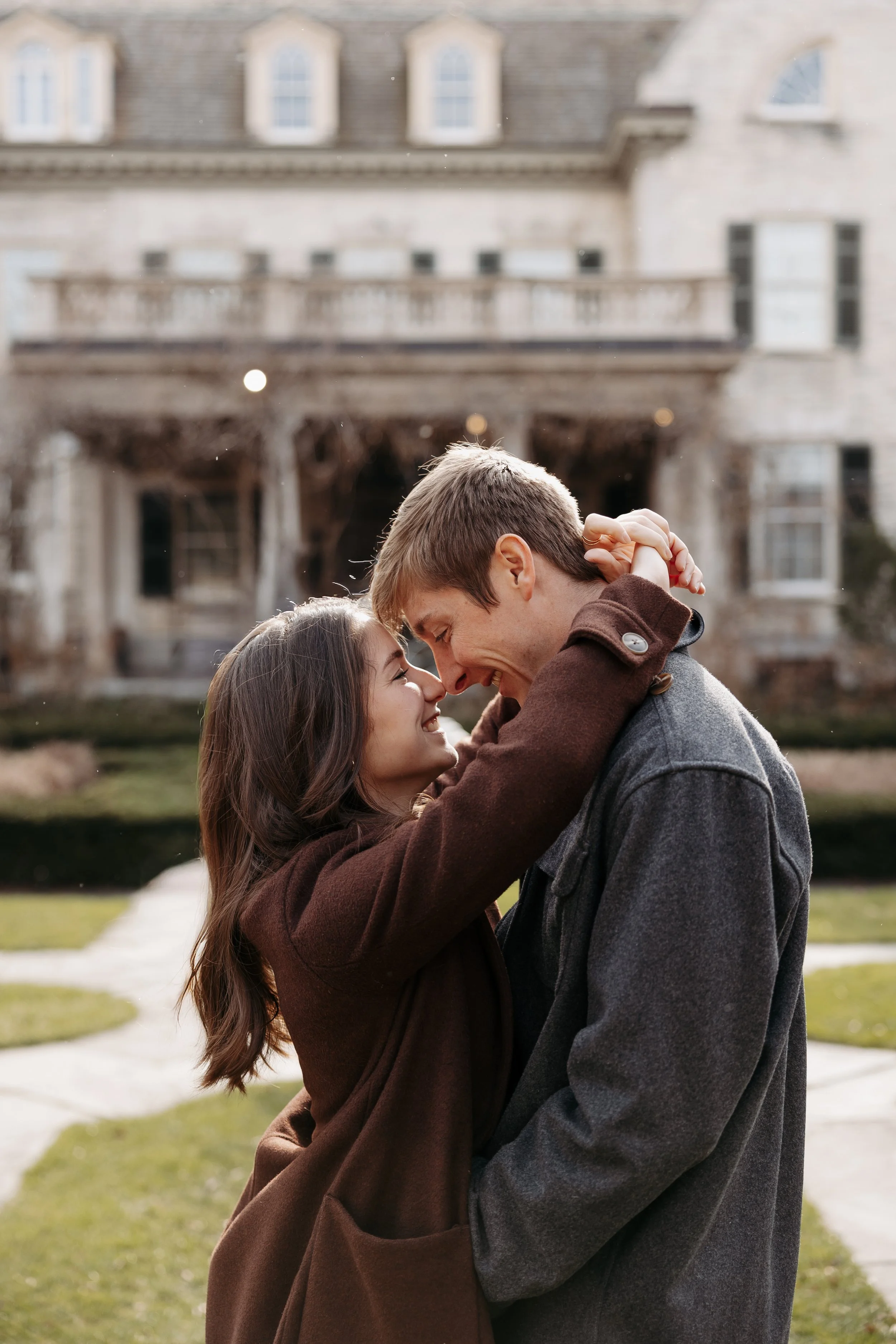 A young couple is outside, embracing and smiling with their noses touching, in front of a large house with multiple windows and balconies. They appear happy and close to each other.