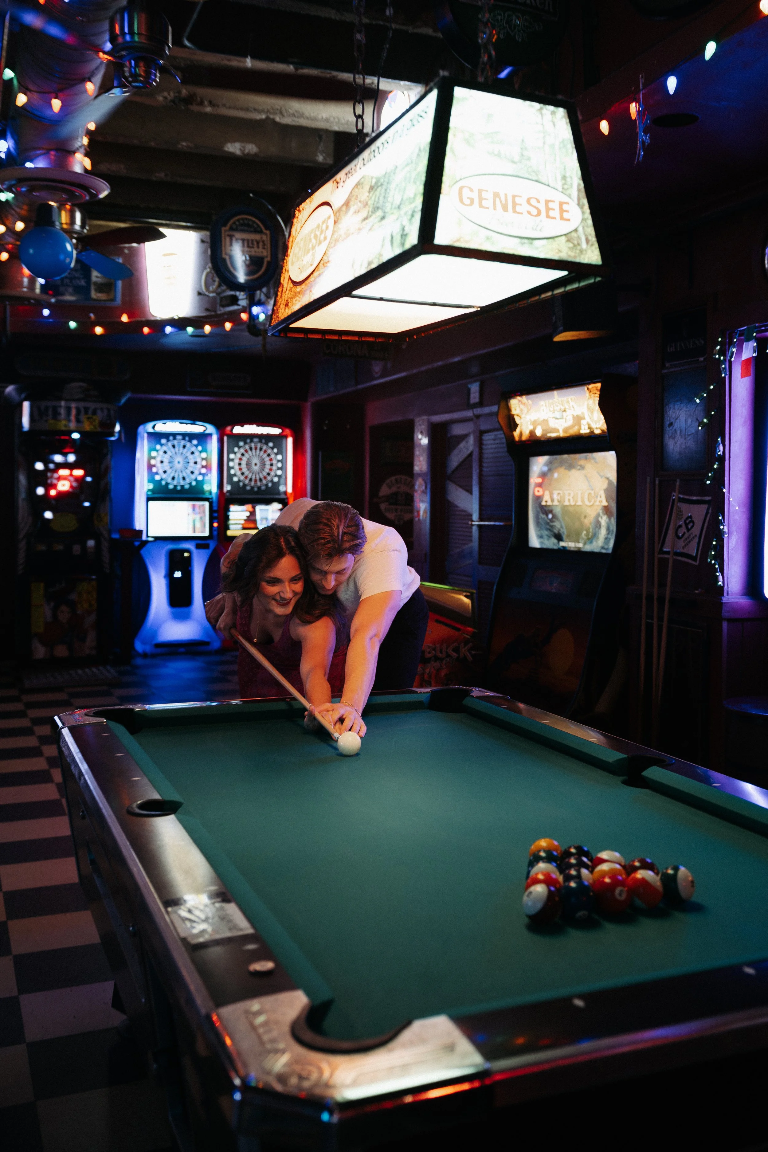 Two people playing pool at a bar with dartboards and TV screens in the background, decorated with string lights and signs.