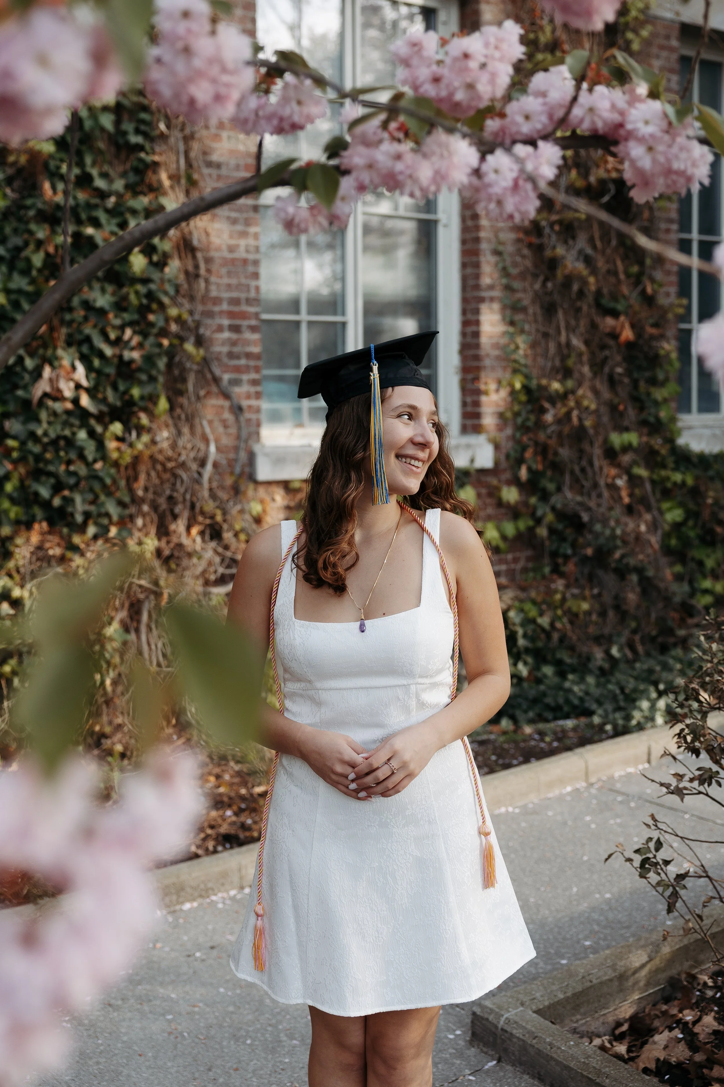 A young woman in a white dress and graduation cap stands outdoors near a pink flowering tree, smiling and looking to her right.