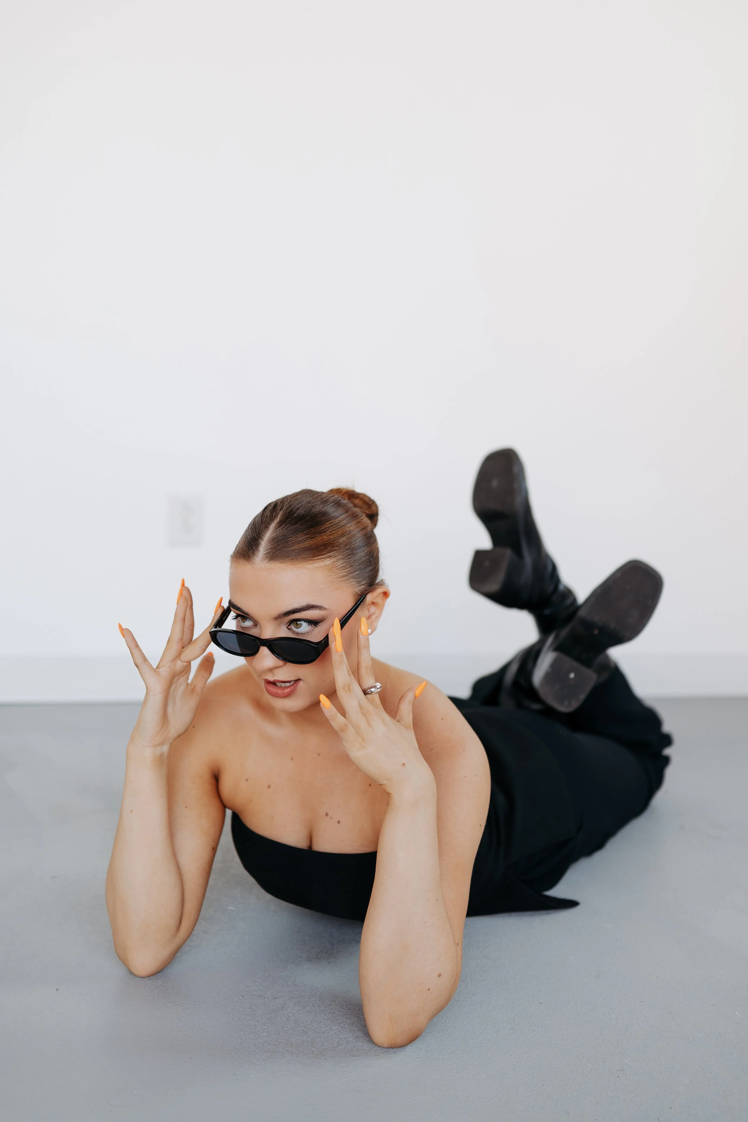 A woman with dark hair tied back, wearing sunglasses, lying on her stomach on the floor with her legs bent at the knees and feet in the air. She is also wearing black clothing and black boots, with a curious or surprised expression as she looks at the camera.