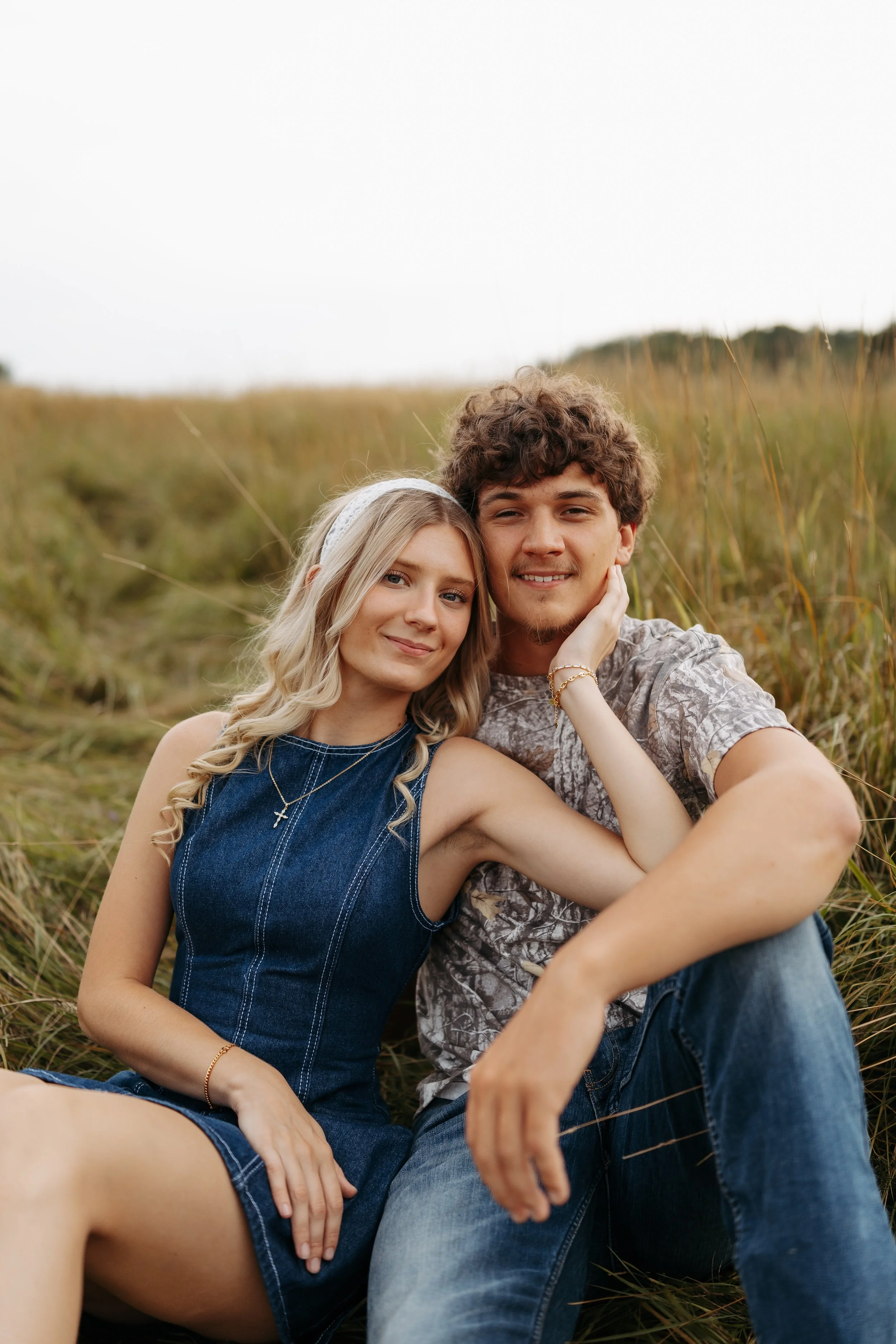 A young couple sitting on the grass in a field, smiling and taking a selfie together. The woman has long blonde hair, is wearing a denim dress and a headband. The man has curly brown hair, is wearing a patterned t-shirt and jeans.
