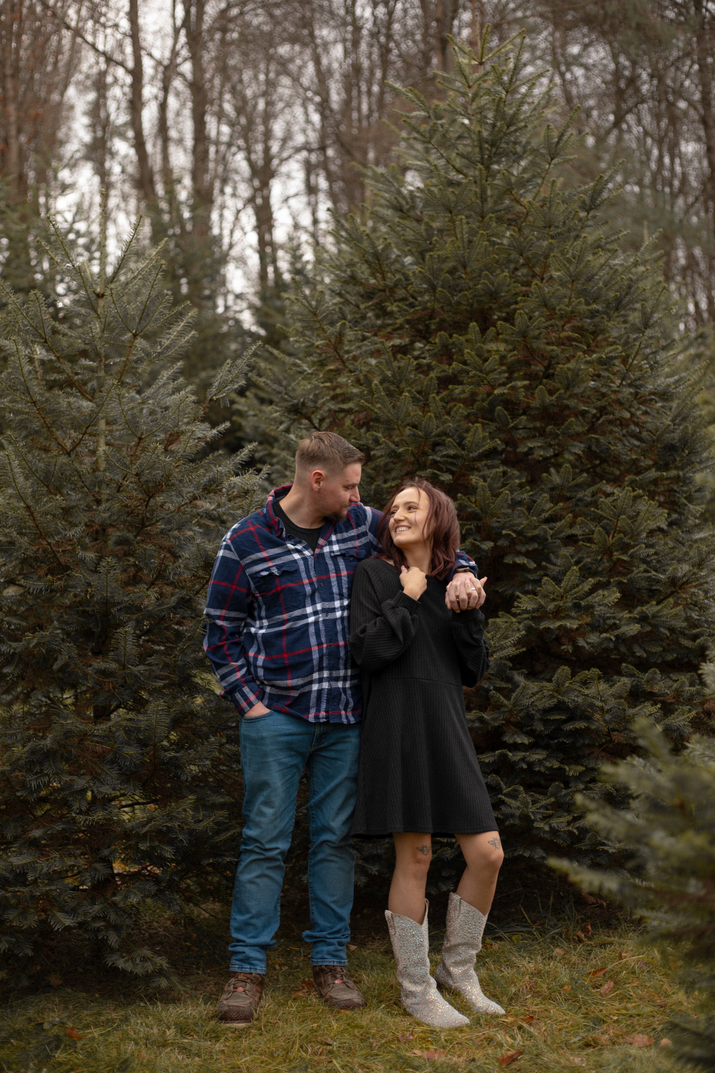 A couple stands among Christmas trees in a forest, smiling and looking at each other, with the woman wearing sparkly boots and the man wearing jeans and a plaid shirt.