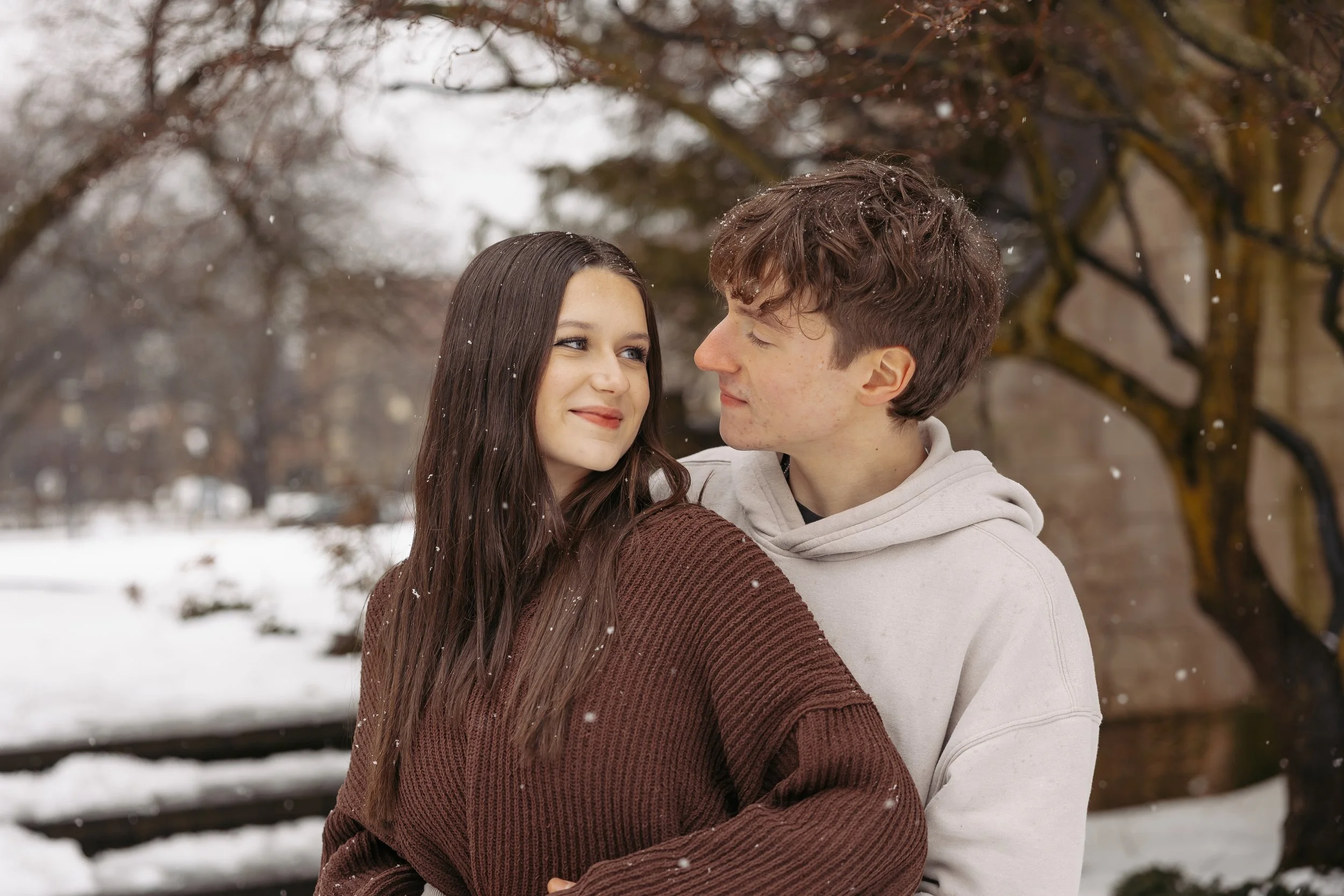 A young man and woman are hugging and smiling at each other outdoors on a snowy winter day, with trees in the background.