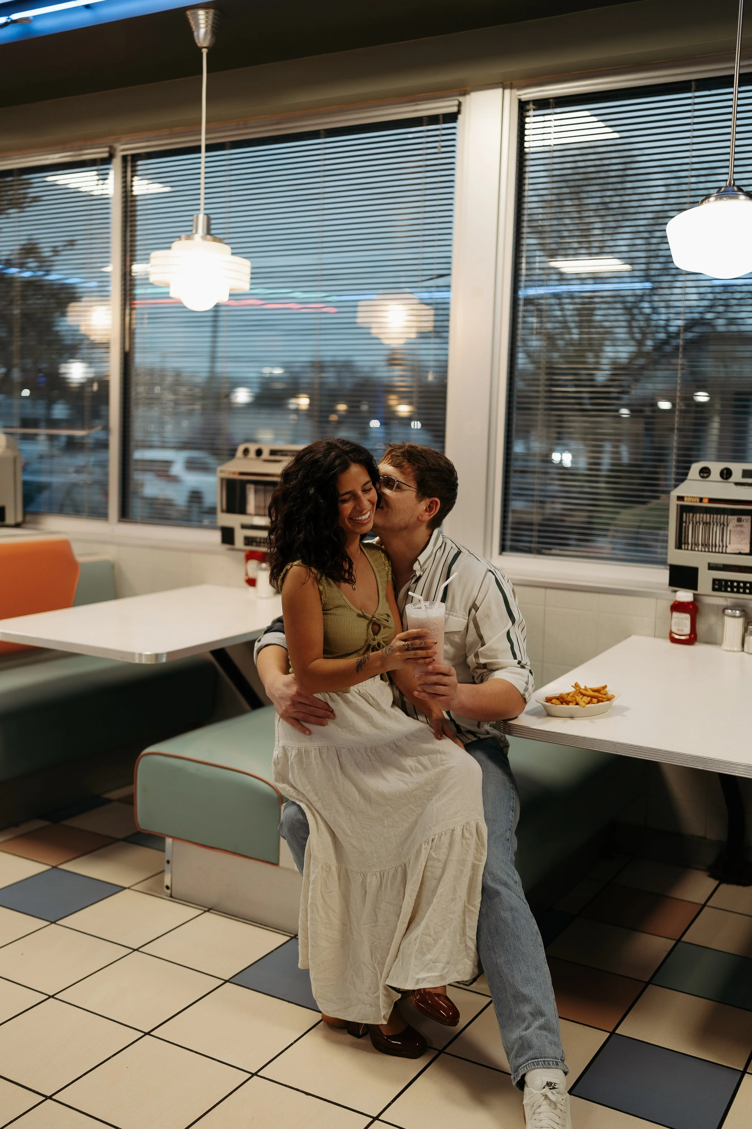 A couple sitting on a diner bench, with the man kissing the woman's cheek, inside a vintage-style American diner with large windows, tiled floor, and pendant lights, while the woman holds a milkshake and there is a plate of fries on the table.