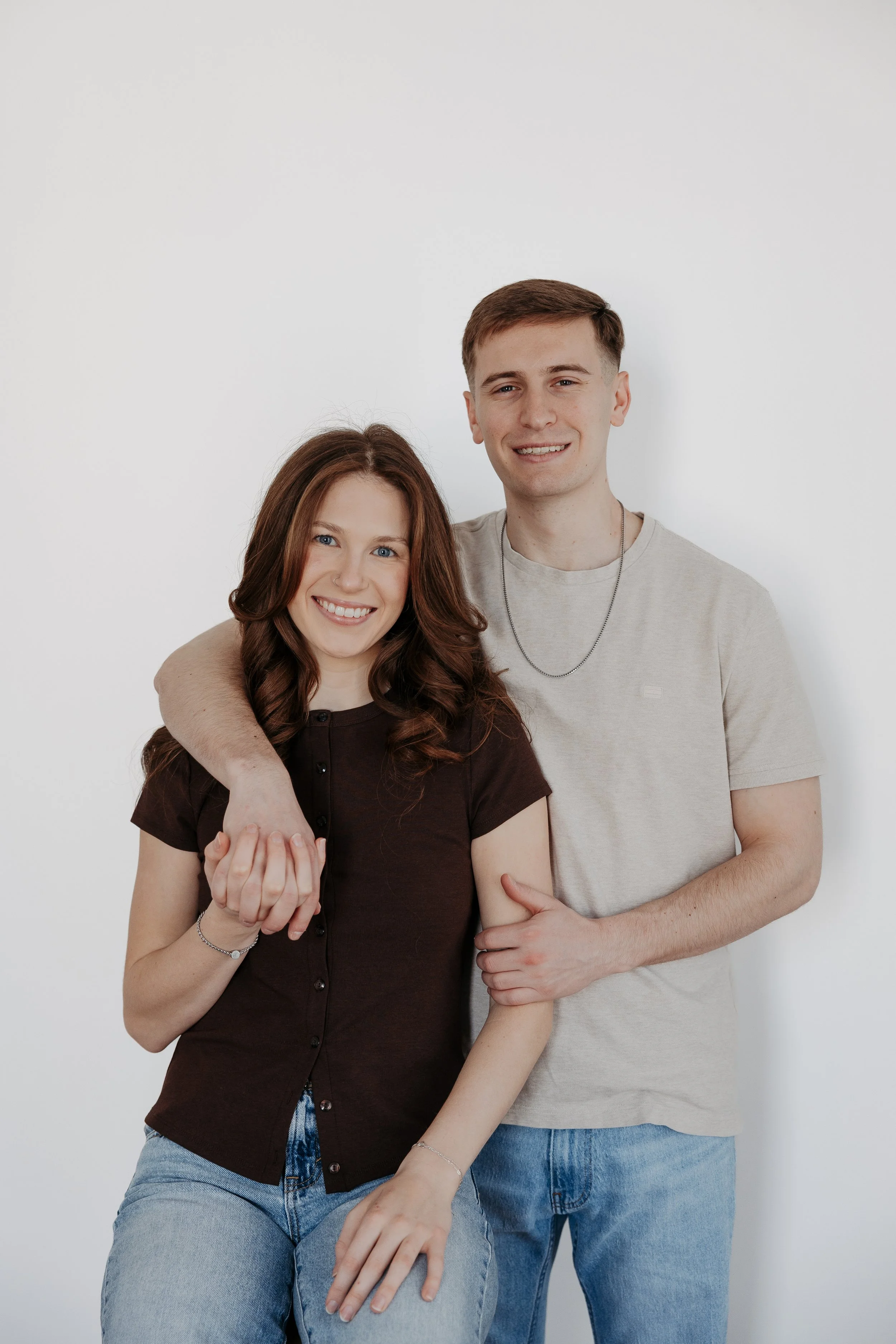 A happy young woman with long brown hair and blue eyes embracing a smiling young man with short light brown hair, both standing against a plain white wall.
