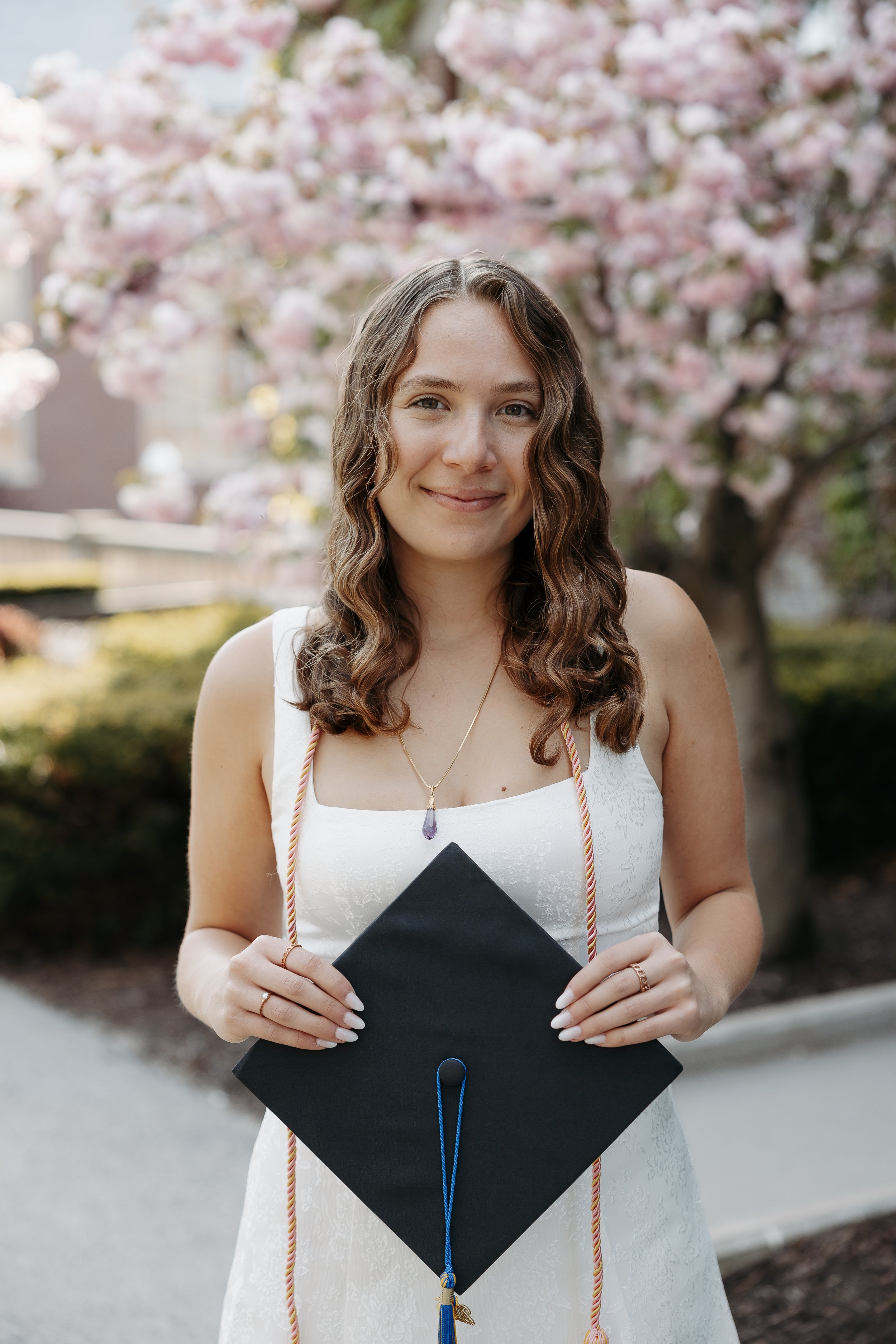 Young woman in a white dress holding a graduation cap, with pink flowering trees in the background.