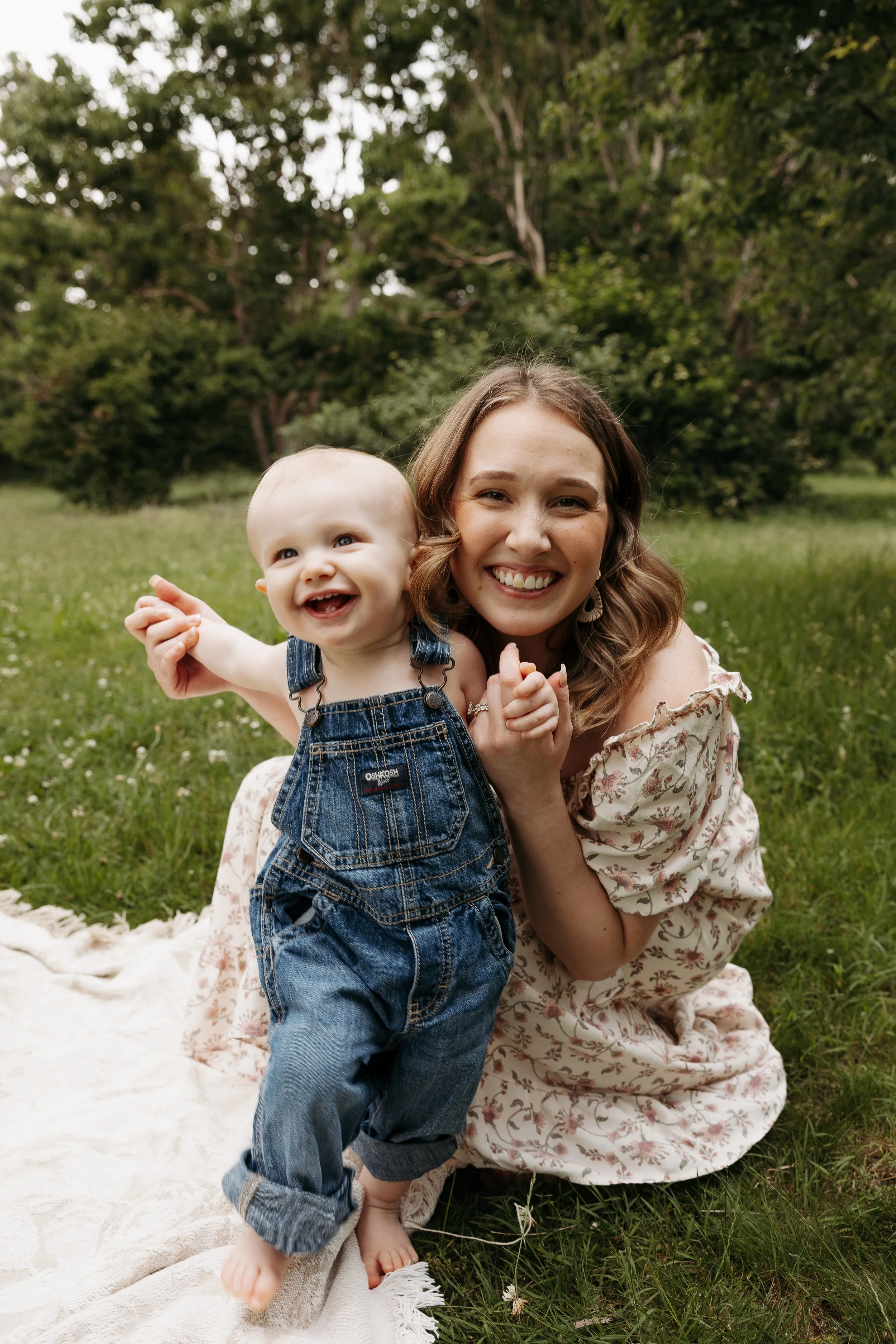 A smiling woman and a happy toddler girl in overalls sitting on a blanket in a grassy park with trees in the background.