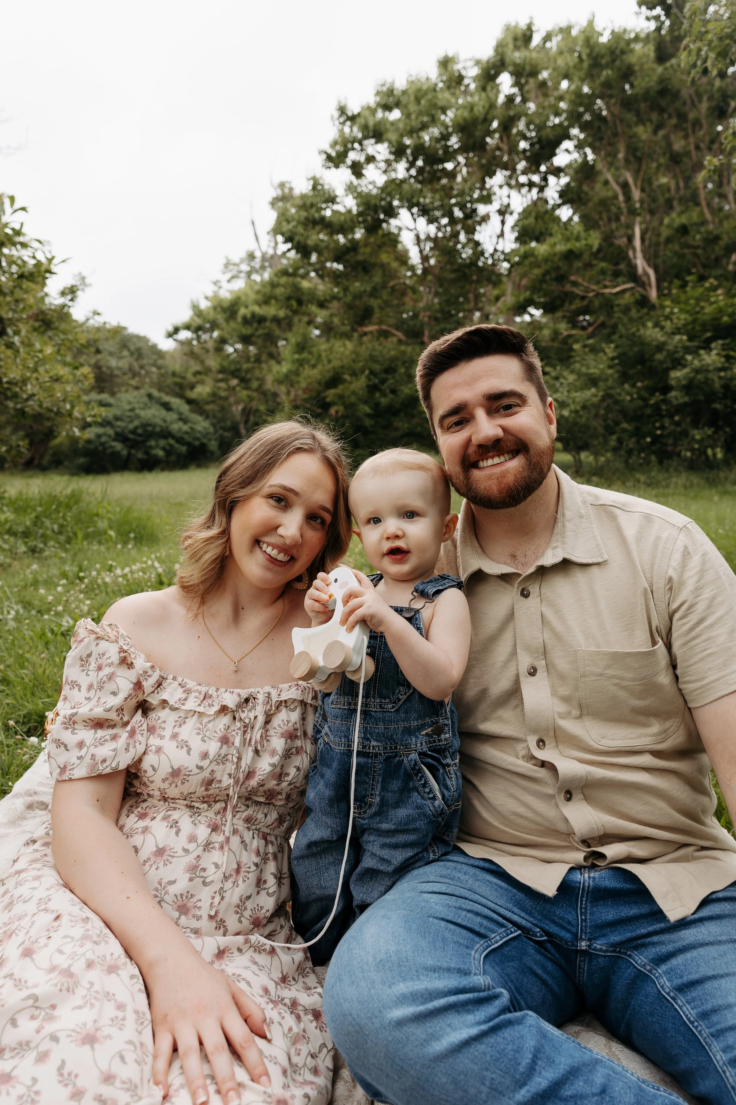 A family of three: a woman with shoulder-length blonde hair, a man with short dark hair and a beard, and a young girl with blonde hair, sitting outdoors in a grassy area with trees in the background. The woman is wearing a floral dress, the man is wearing a beige shirt, and the girl is holding a wooden toy with balloons.