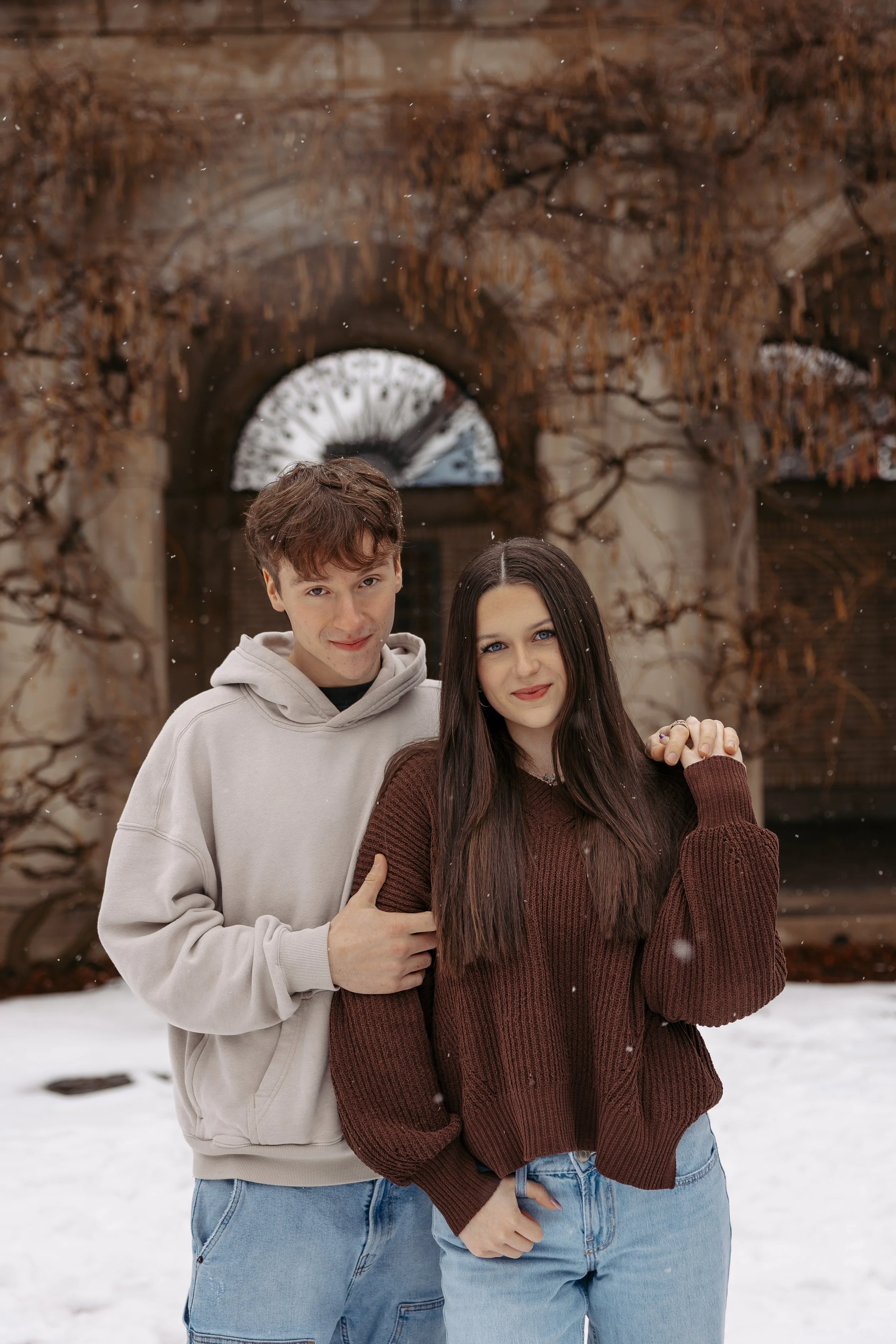A young man and woman standing close together outside in the snow, holding hands, in front of a brick building with bare tree branches. The man wears a light hoodie, and the woman wears a brown sweater.