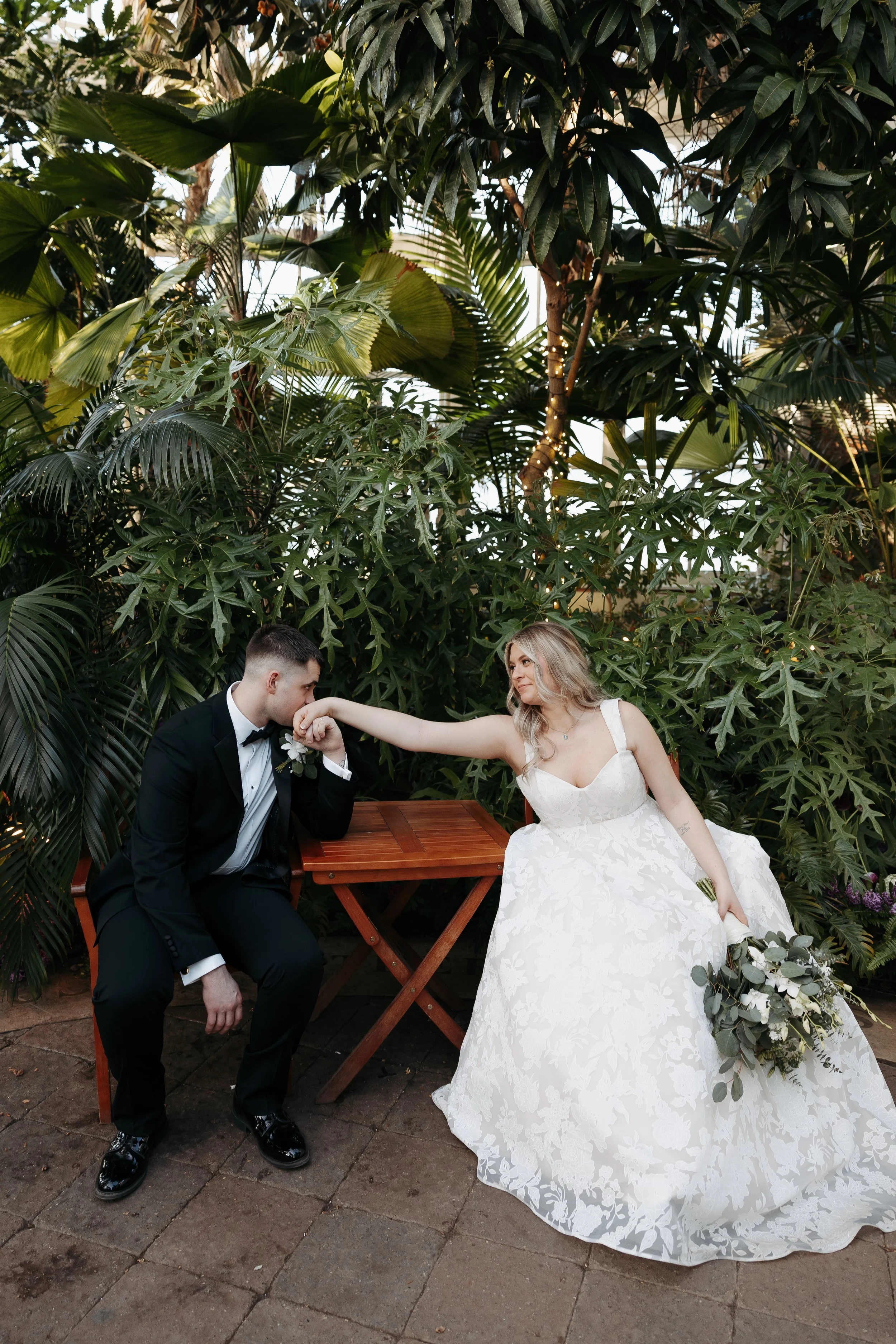 A bride and groom are sitting outdoors in a lush green garden. The groom is dressed in a black tuxedo and bow tie, kneeling with his right knee on the ground and holding the bride's hand. The bride is wearing a white wedding gown with floral patterns
