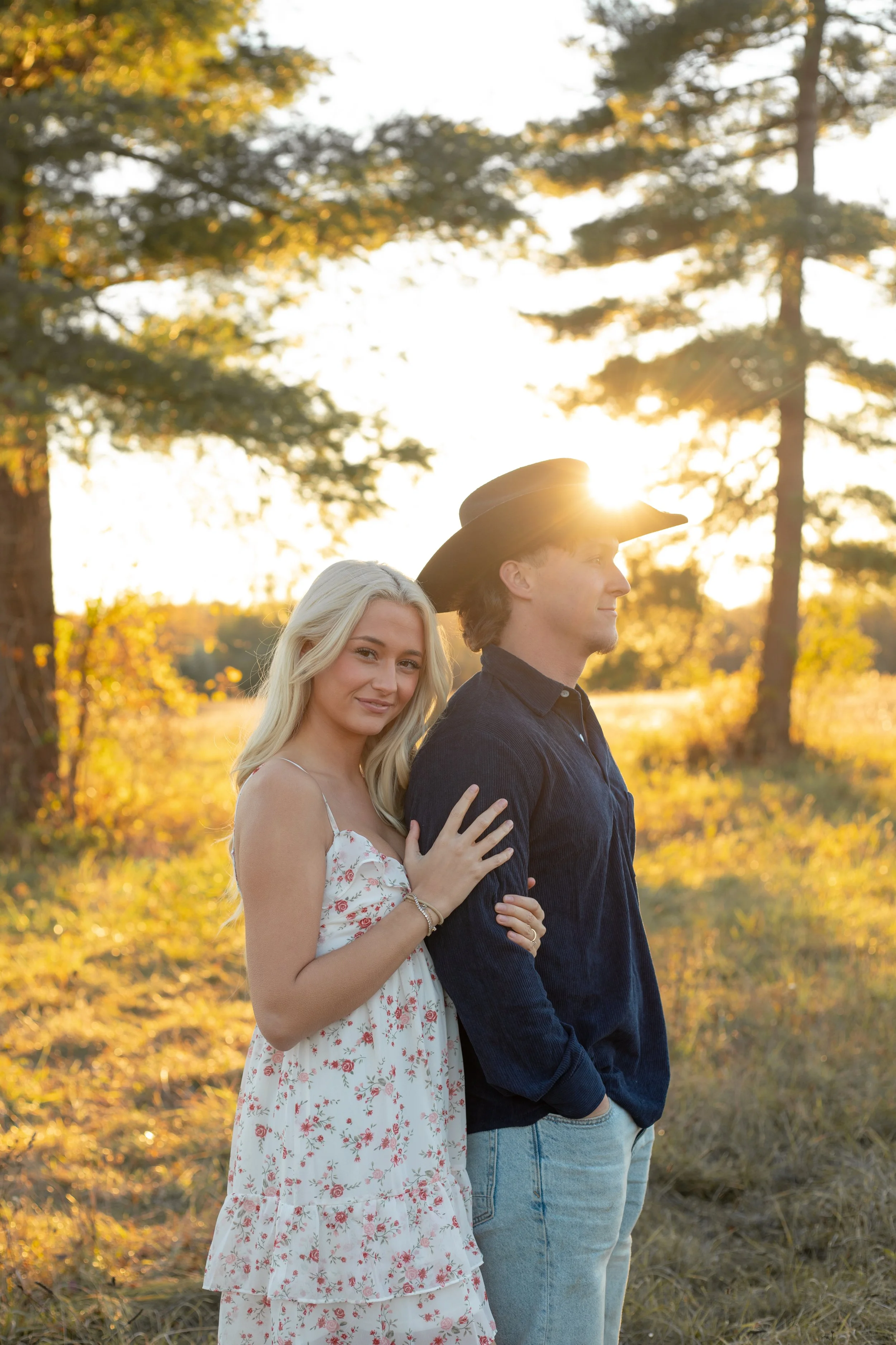 A young woman with blonde hair and a young man with a cowboy hat outdoors during sunset, standing in a grassy area with trees in the background.