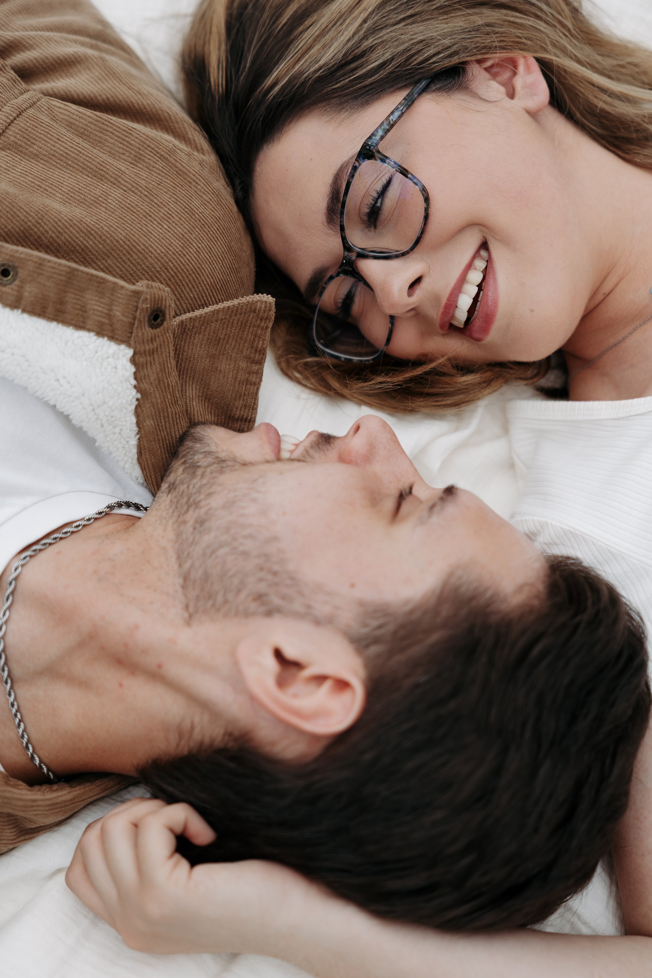 A smiling woman with glasses lying close to a man with dark hair, both resting their heads on a white surface, appearing happy and relaxed.