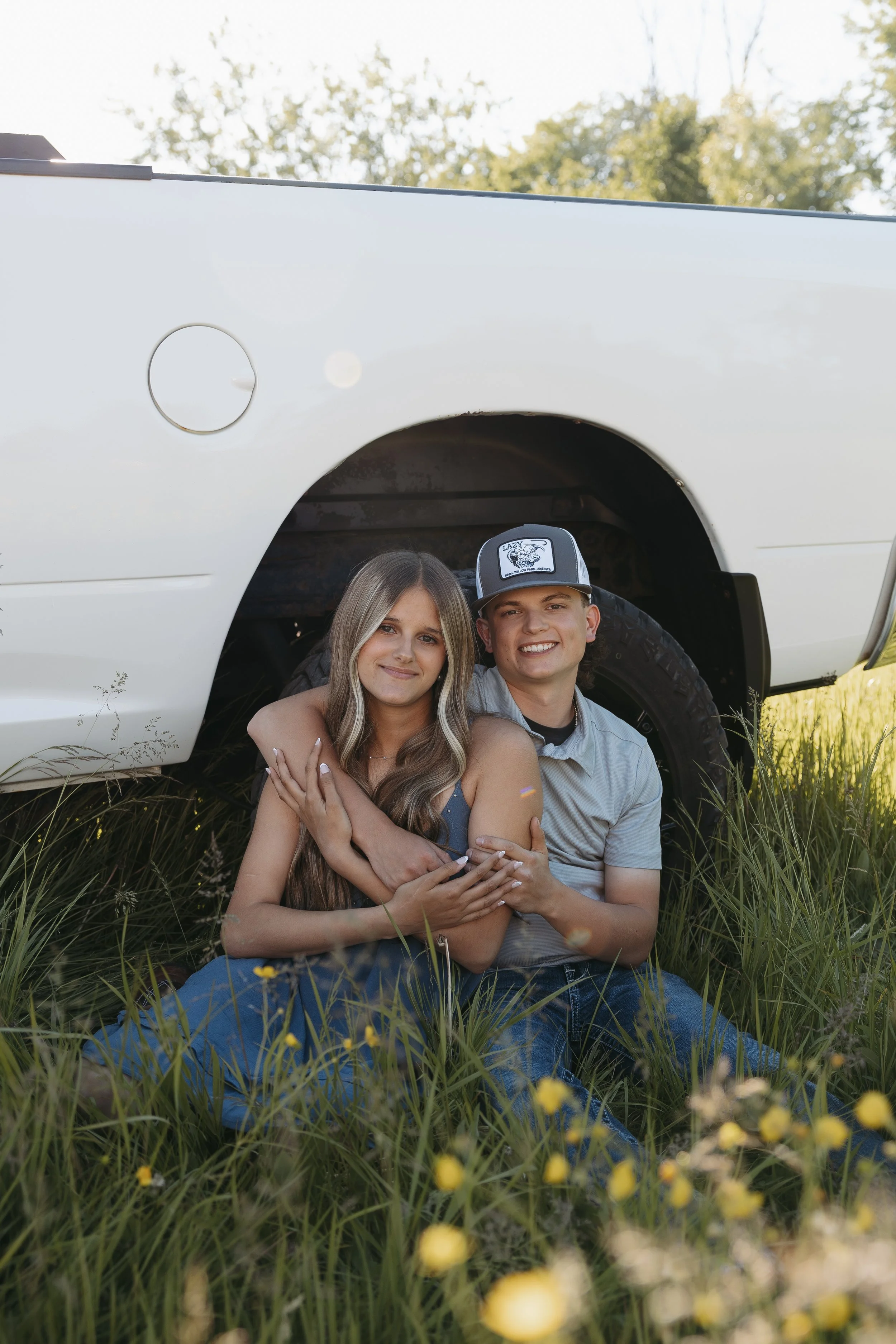 A young couple sitting on grass in front of a white vehicle, with a woman with long hair and a man wearing a baseball cap, smiling and embracing each other outdoors.
